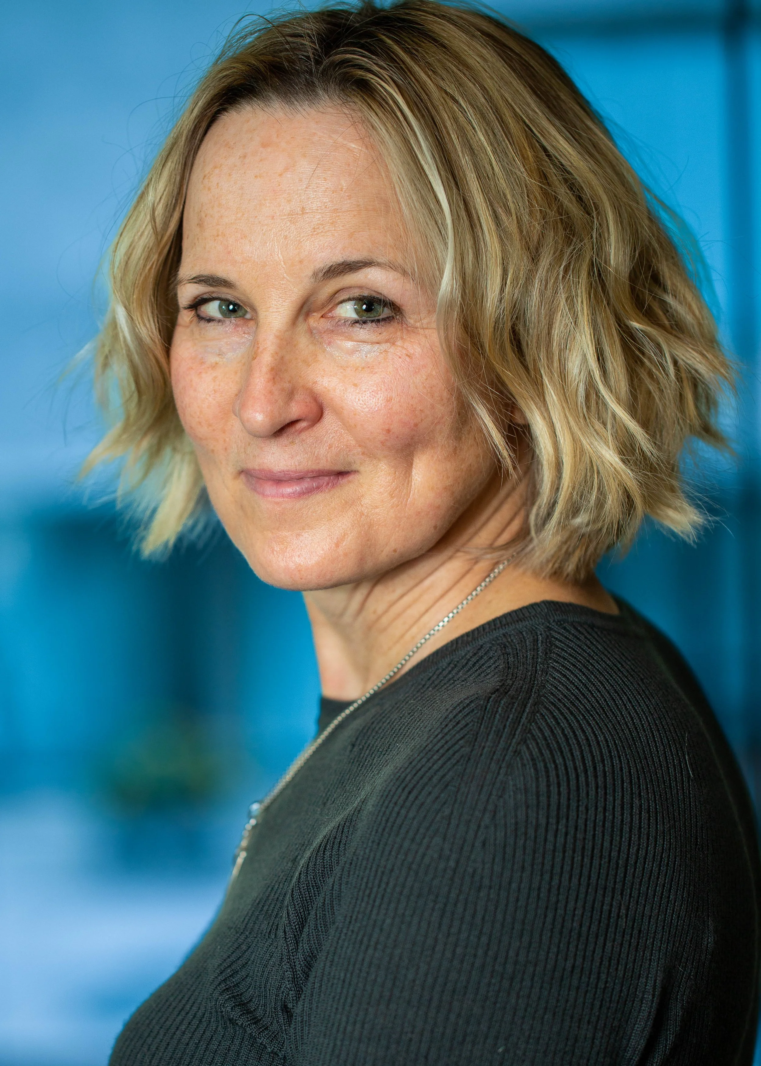 A middle-aged woman with shoulder-length blonde hair smiling at the camera, wearing a black top and a silver necklace, against a blue blurred background.