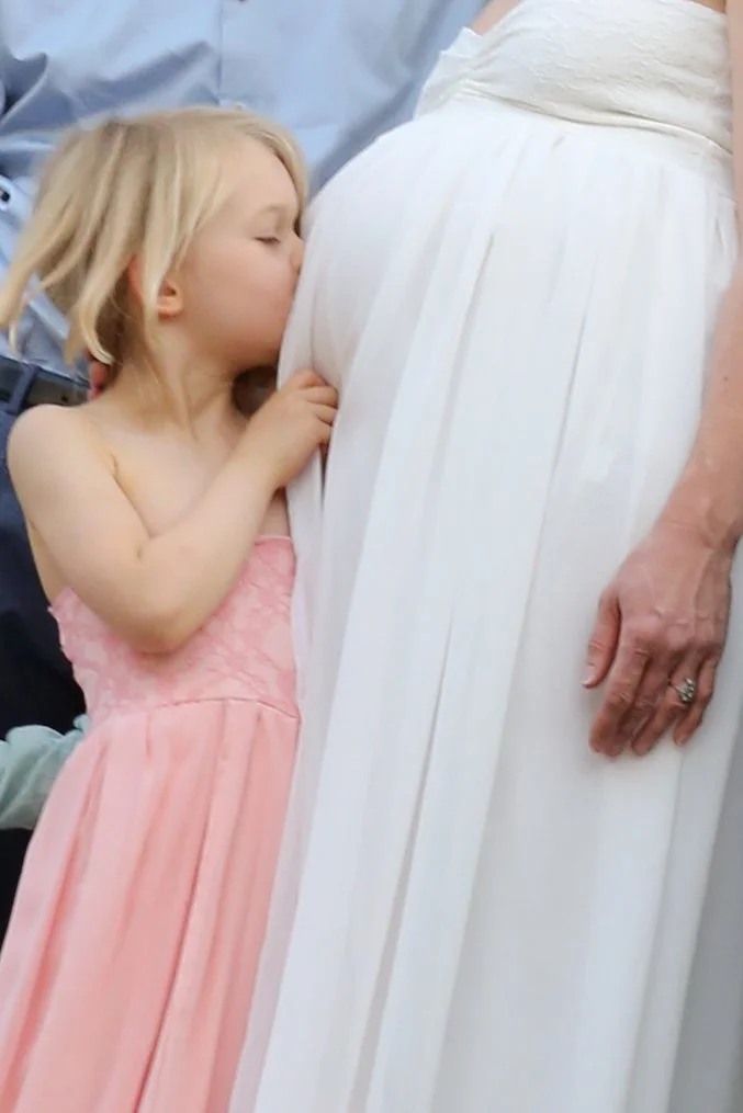 A young girl in a pink dress kisses a pregnant woman's belly during a special moment.