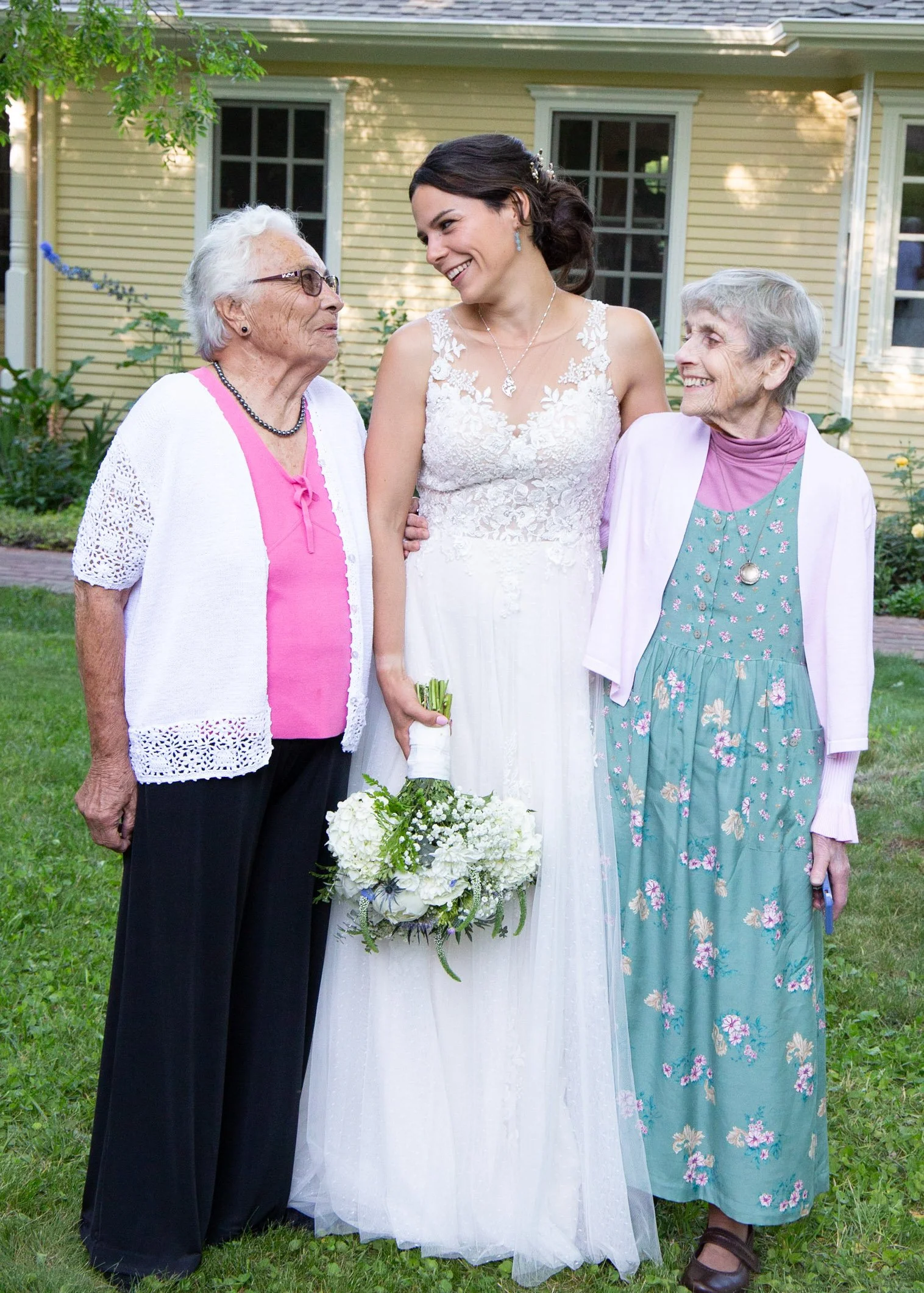 A bride in a white wedding dress holding a bouquet, standing outdoors with two elderly women, all smiling and facing each other, in front of a yellow house with green lawn.