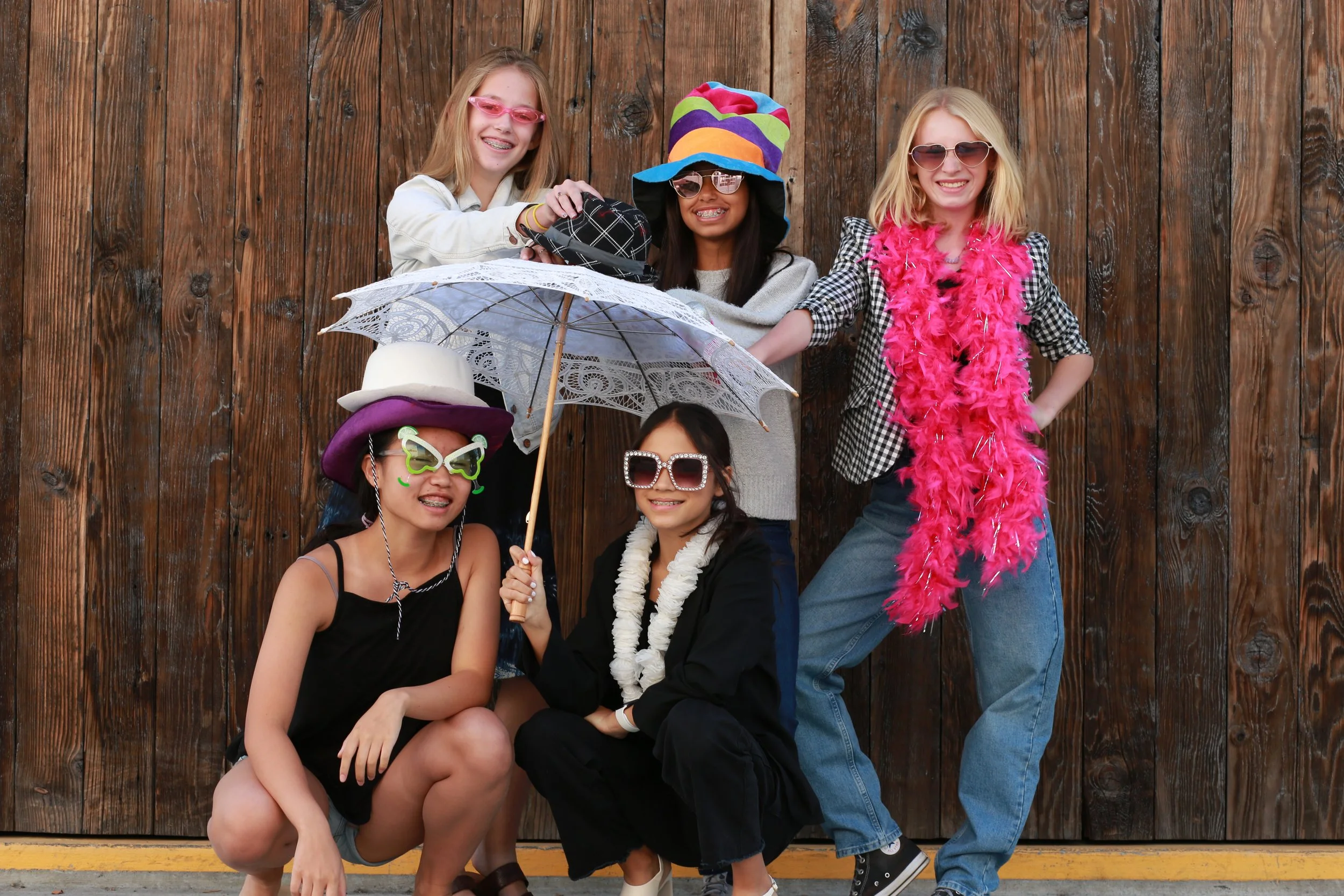 A group of six girls dressed in colorful, playful outfits posing in front of a wooden wall. They are wearing sunglasses, hats, feather boas, and holding umbrellas, smiling and having fun.