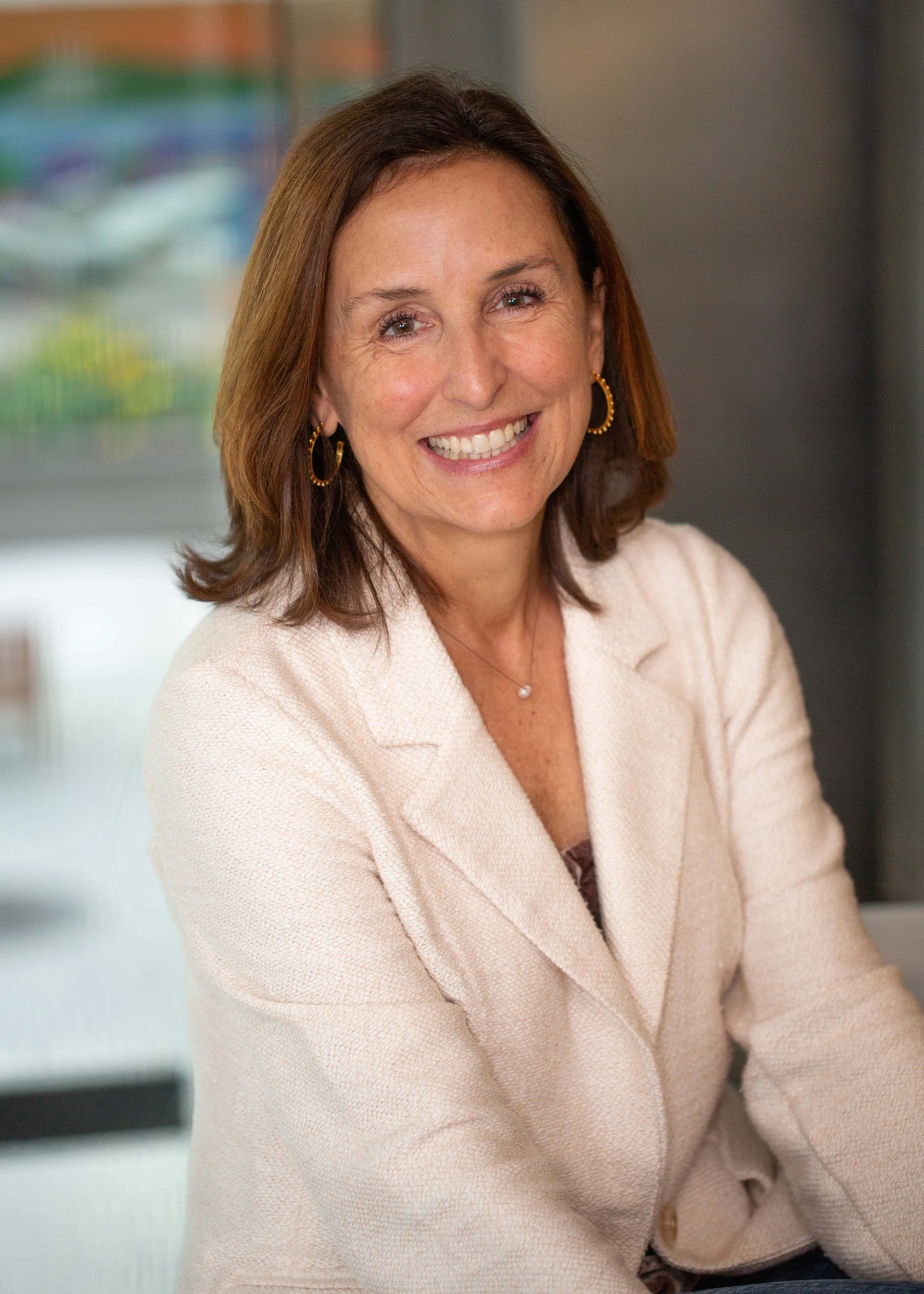 A middle-aged woman with shoulder-length brown hair, wearing a cream-colored blazer, earrings, and a necklace, smiling at the camera with an office background.