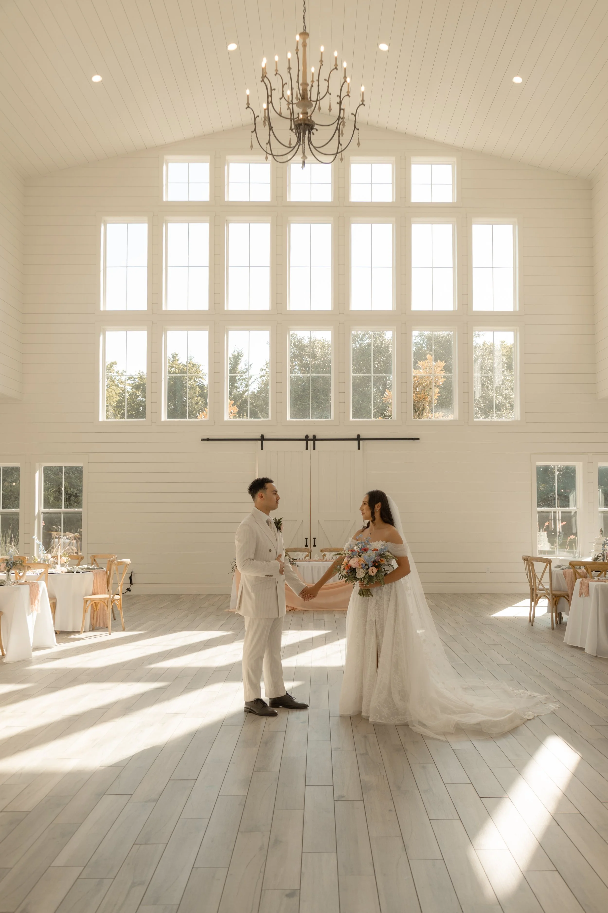 A bride and groom stand holding hands in a bright, spacious wedding venue with large windows and a high, vaulted ceiling. The bride wears a long white gown and holds a bouquet, while the groom wears a light-colored suit and tie. Sunlight streams through the windows, illuminating the scene.