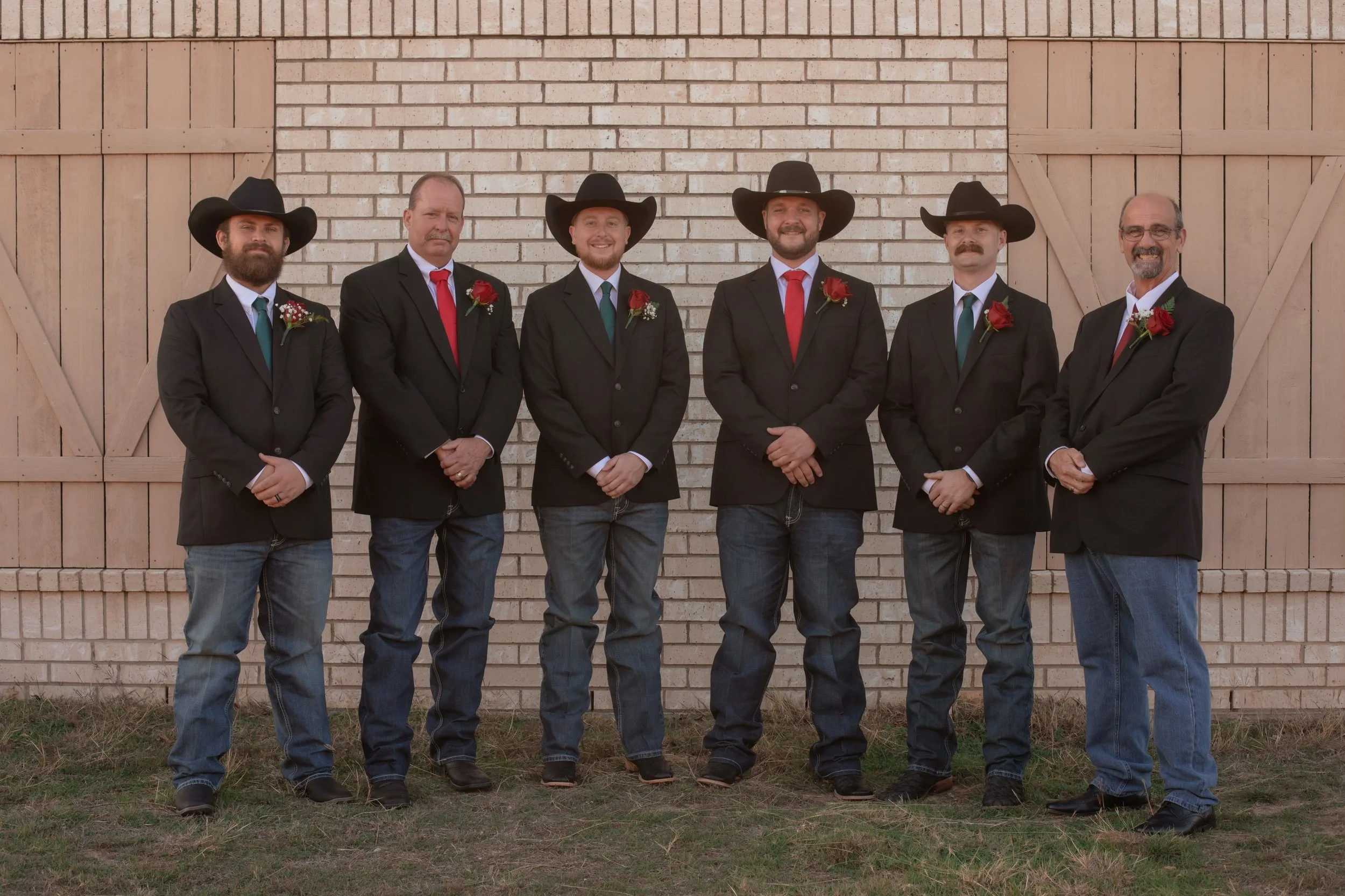 Group of six men in formal black suits, jeans, and cowboy hats standing in front of a brick wall and wooden barn doors, each with a red rose boutonniere.