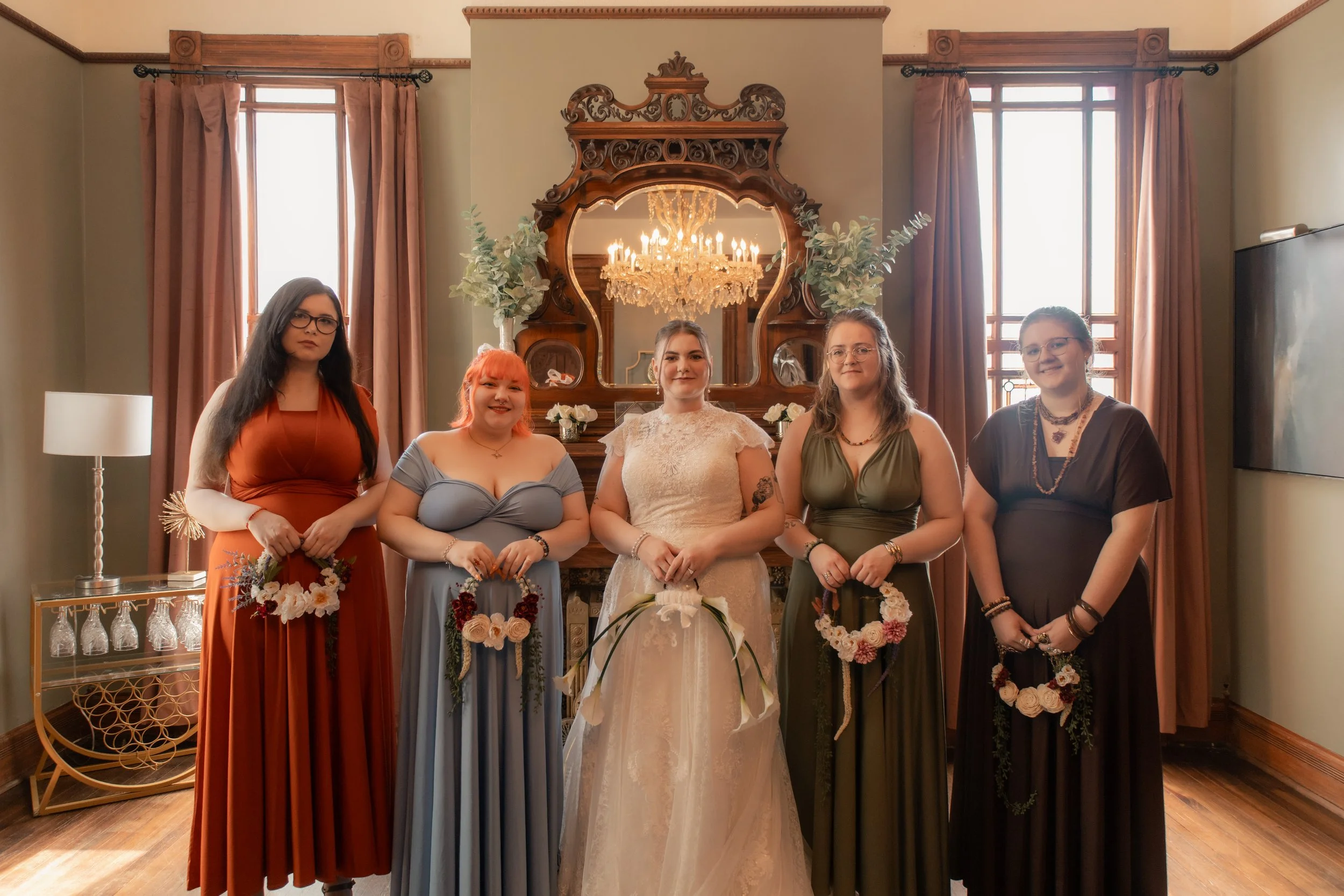 A group of five women standing indoors with a large decorative wooden mirror with a chandelier reflected in it behind them. The women are dressed in colorful dresses, holding floral bouquets, and standing in front of a vintage-style mirror with a cha