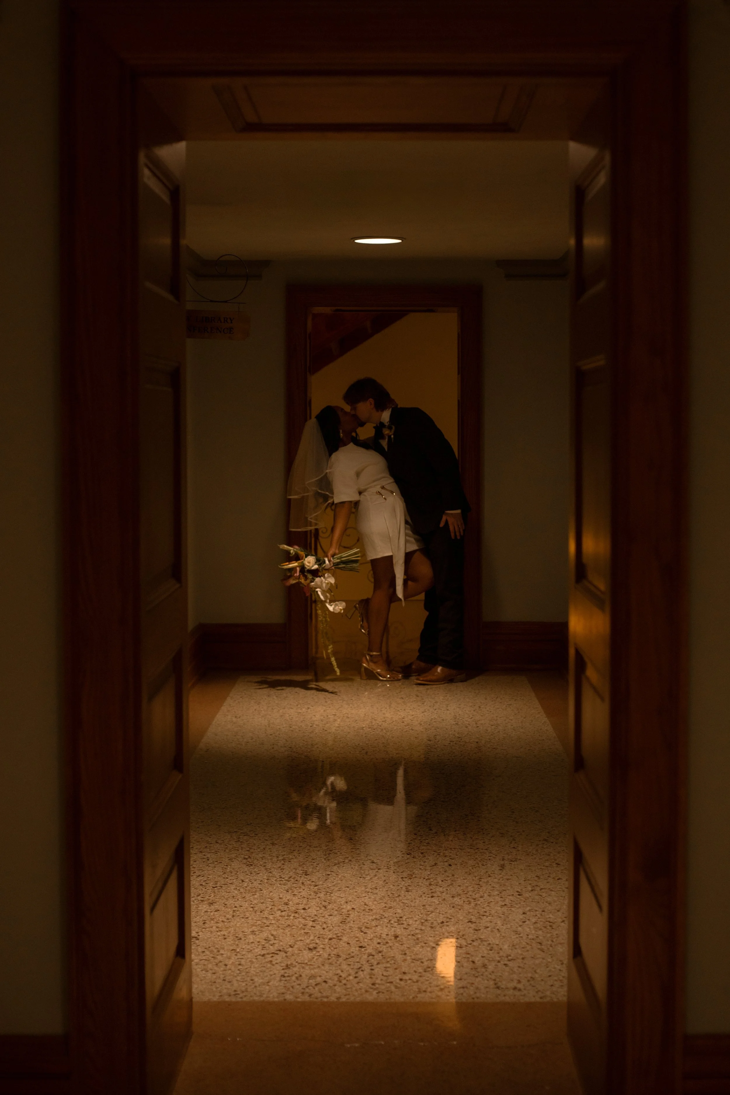 A romantic moment between two people dressed in wedding attire, sharing a kiss in a hallway reflected on the floor.