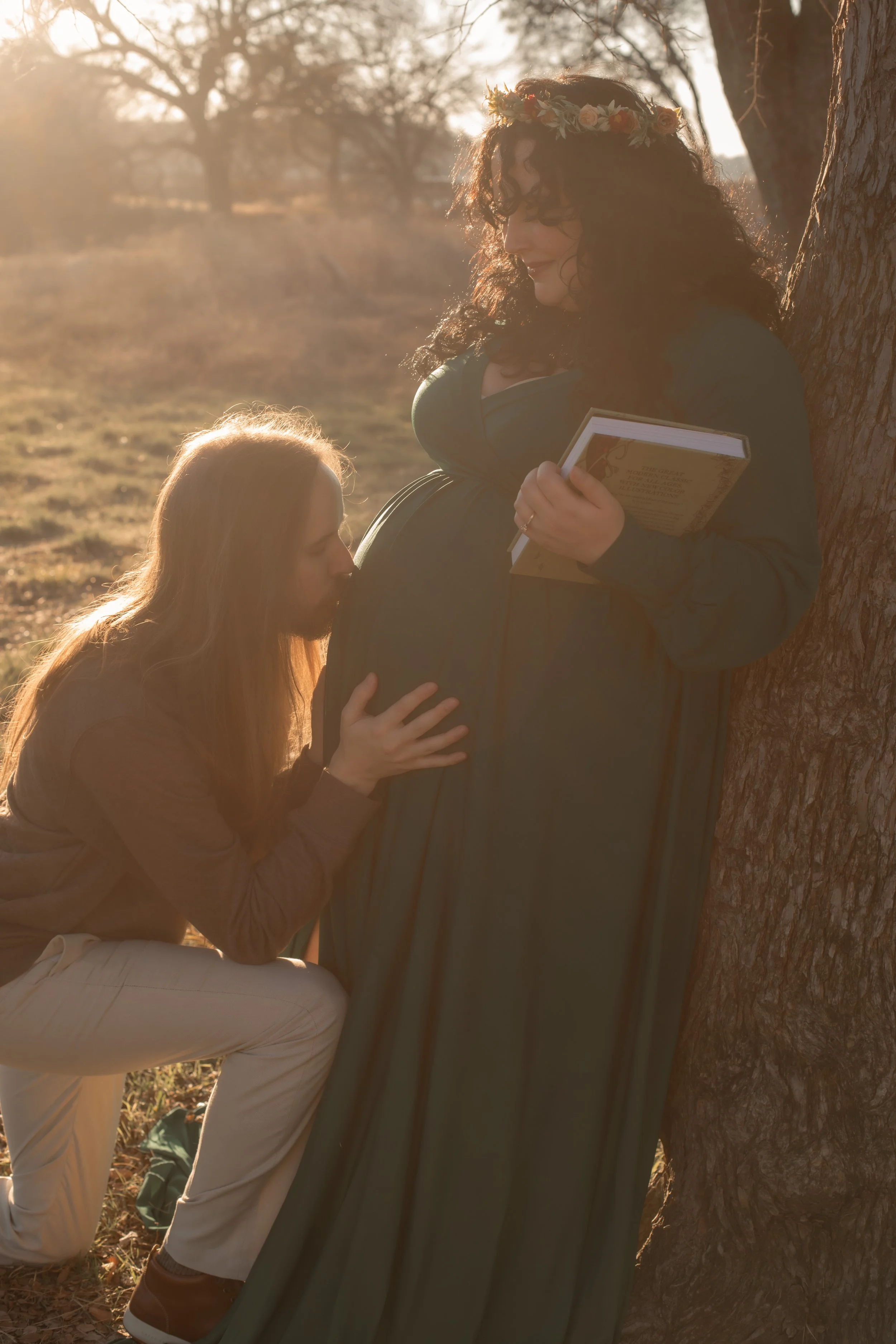 A pregnant woman with curly hair, wearing a flower crown and a dark green dress, stands next to a tree at sunset, holding a book. A man with long hair, wearing a brown jacket and beige pants, is kneeling and kissing her belly, gently holding it.