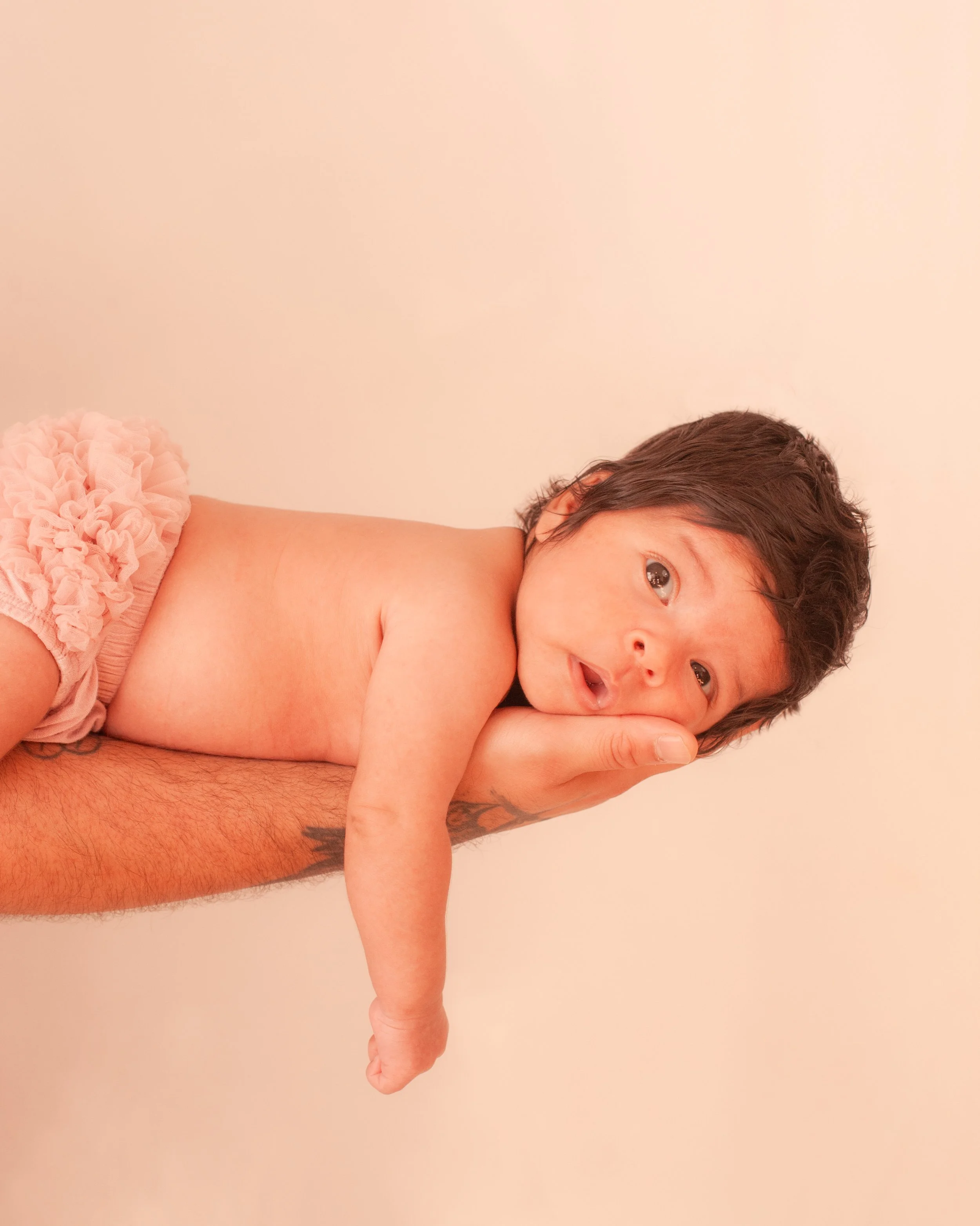 A newborn baby with dark hair lying on an adult's hand against a plain beige background.