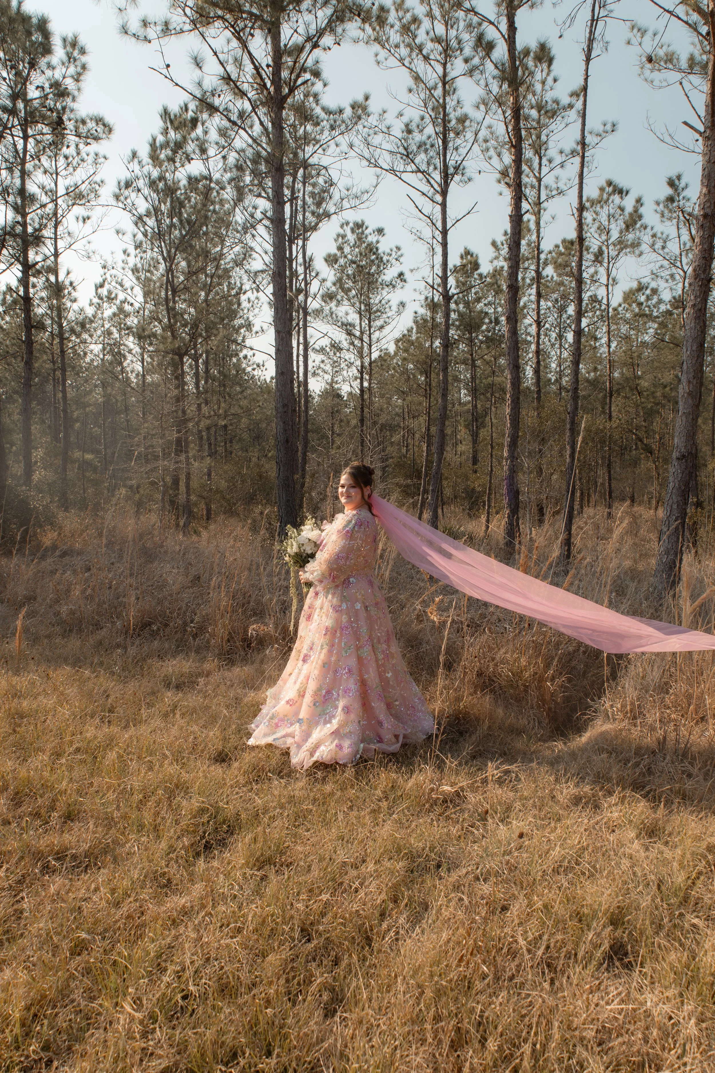 A woman in a floral pink wedding dress holding a bouquet standing in a field of dry grass with tall trees in the background, wearing a pink veil flowing behind her.