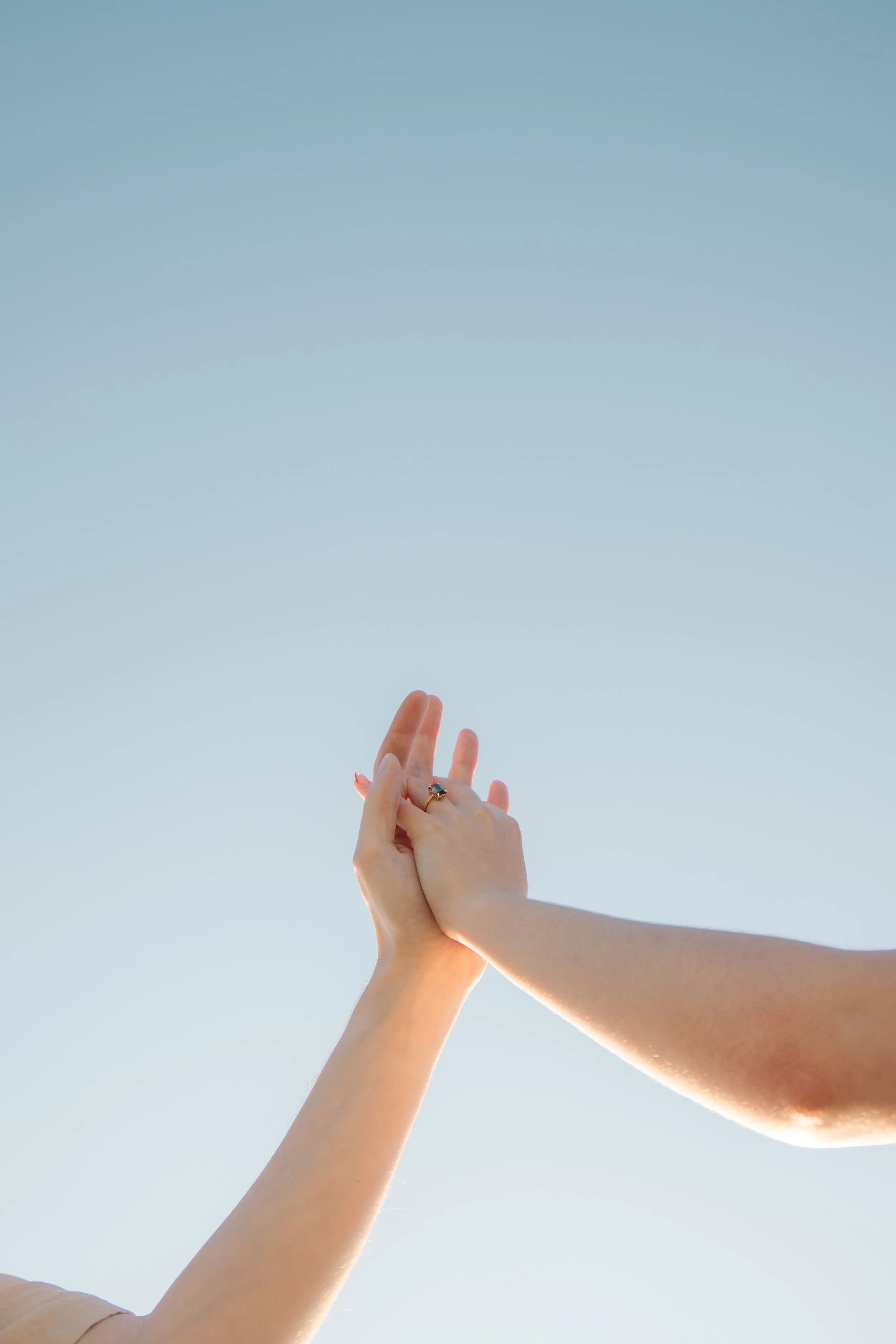 Two hands, one with a ring, touching in the air with a clear sky in the background.