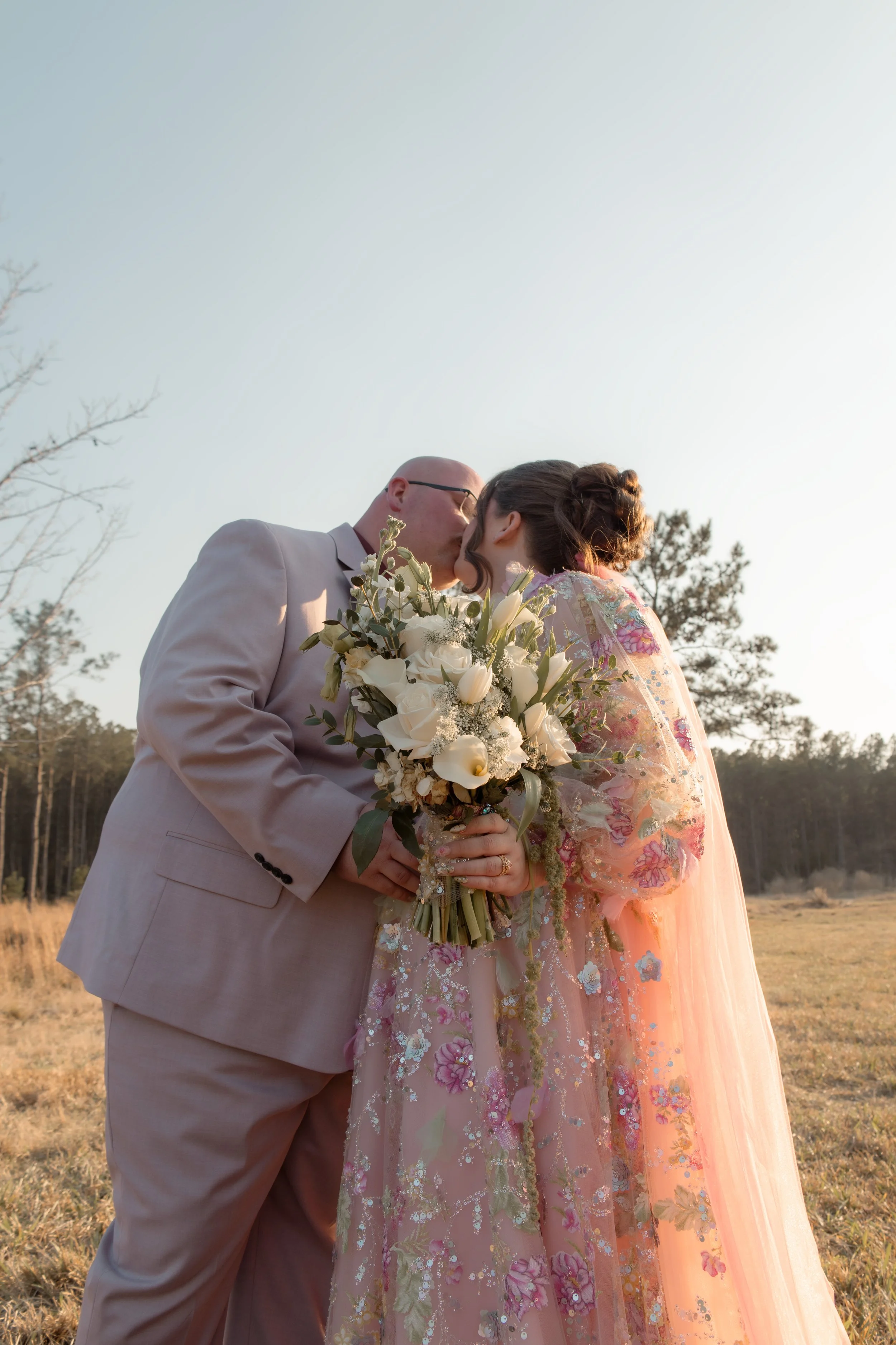 A couple sharing a kiss outdoors, dressed in wedding attire, holding a bouquet of white flowers, with trees and open field in the background during sunset.