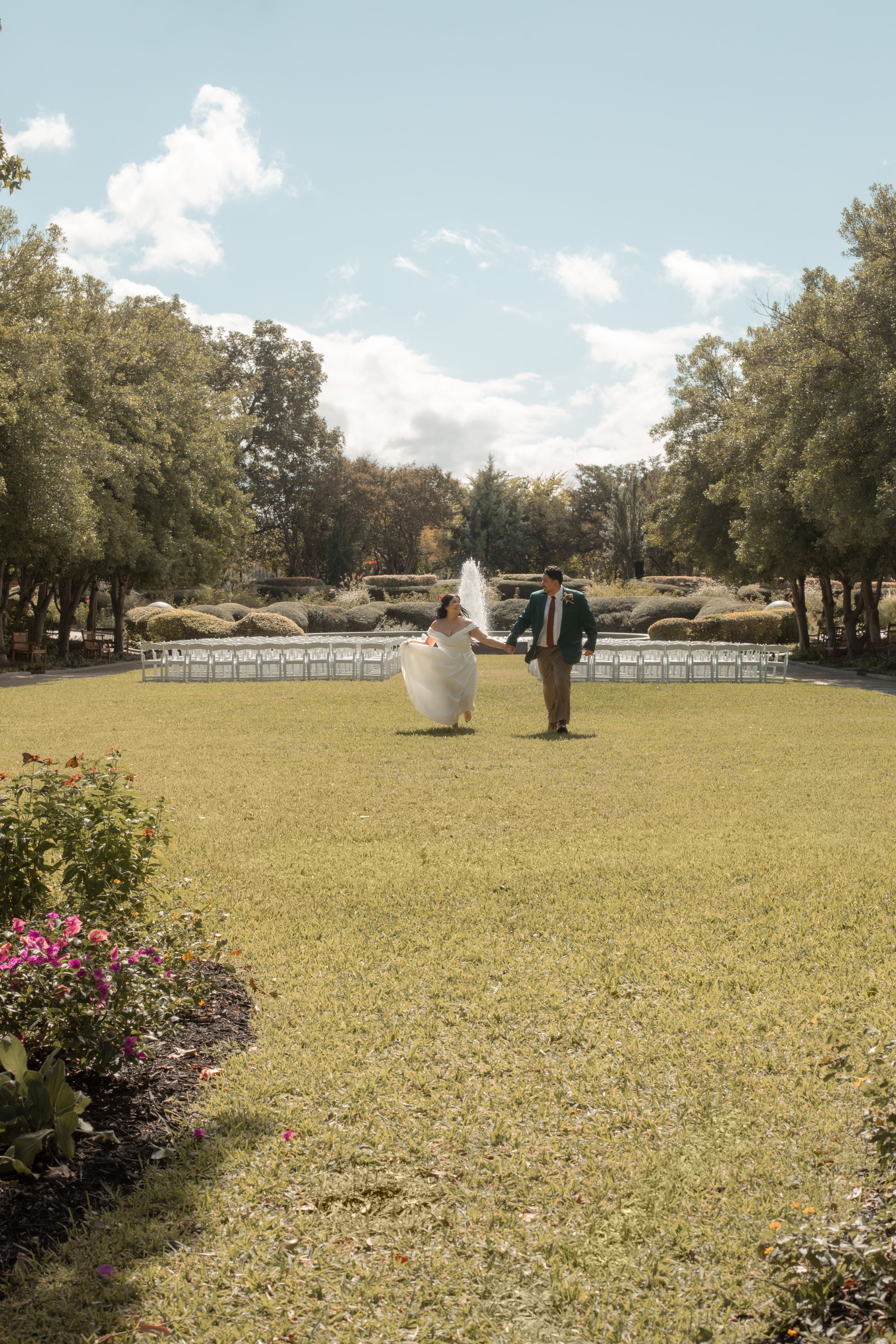 A newlywed couple holding hands and dancing on a grassy lawn in a park with a fountain and trees in the background.