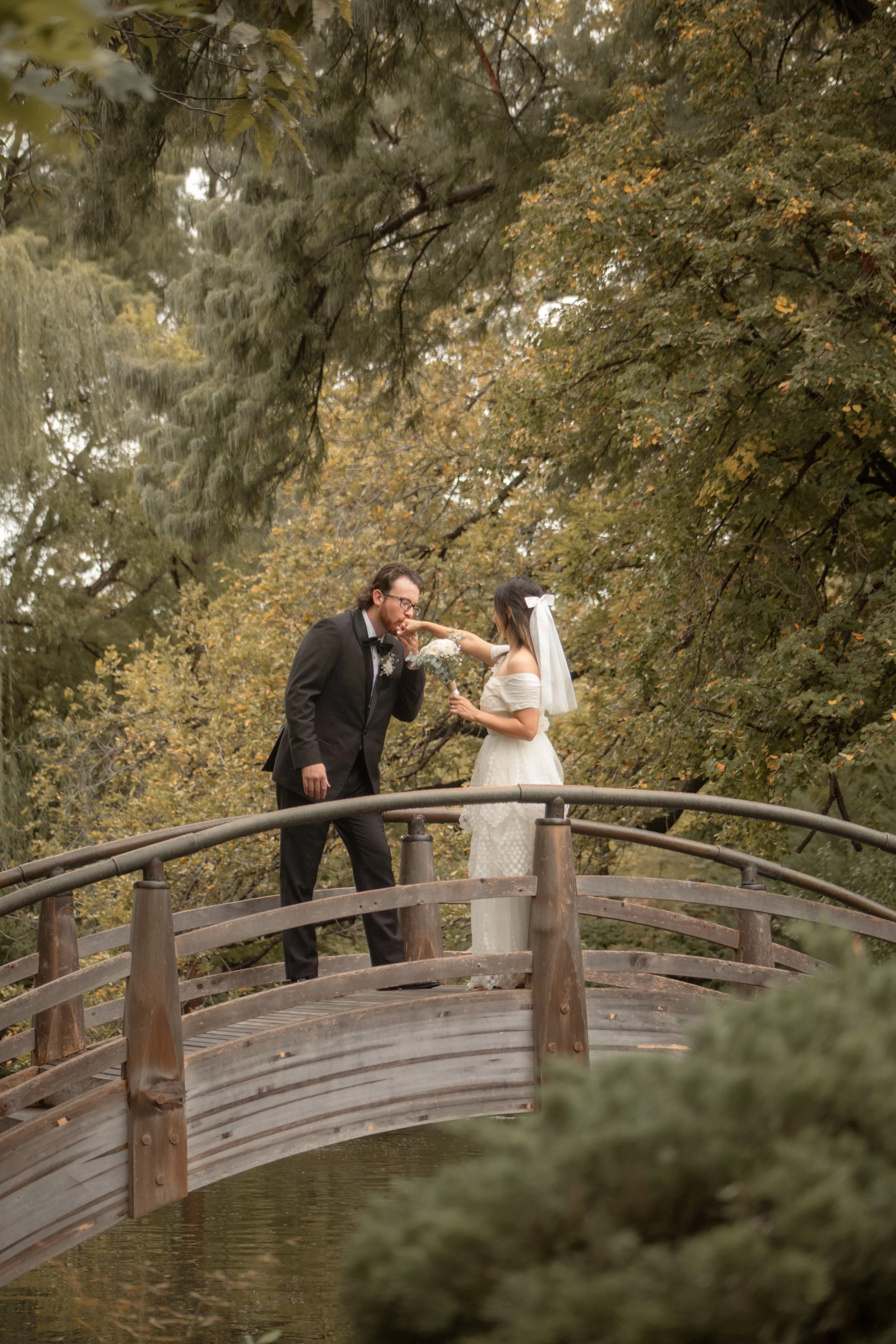 A bride and groom standing on a wooden bridge surrounded by trees, with the groom kissing the bride's hand during their wedding.