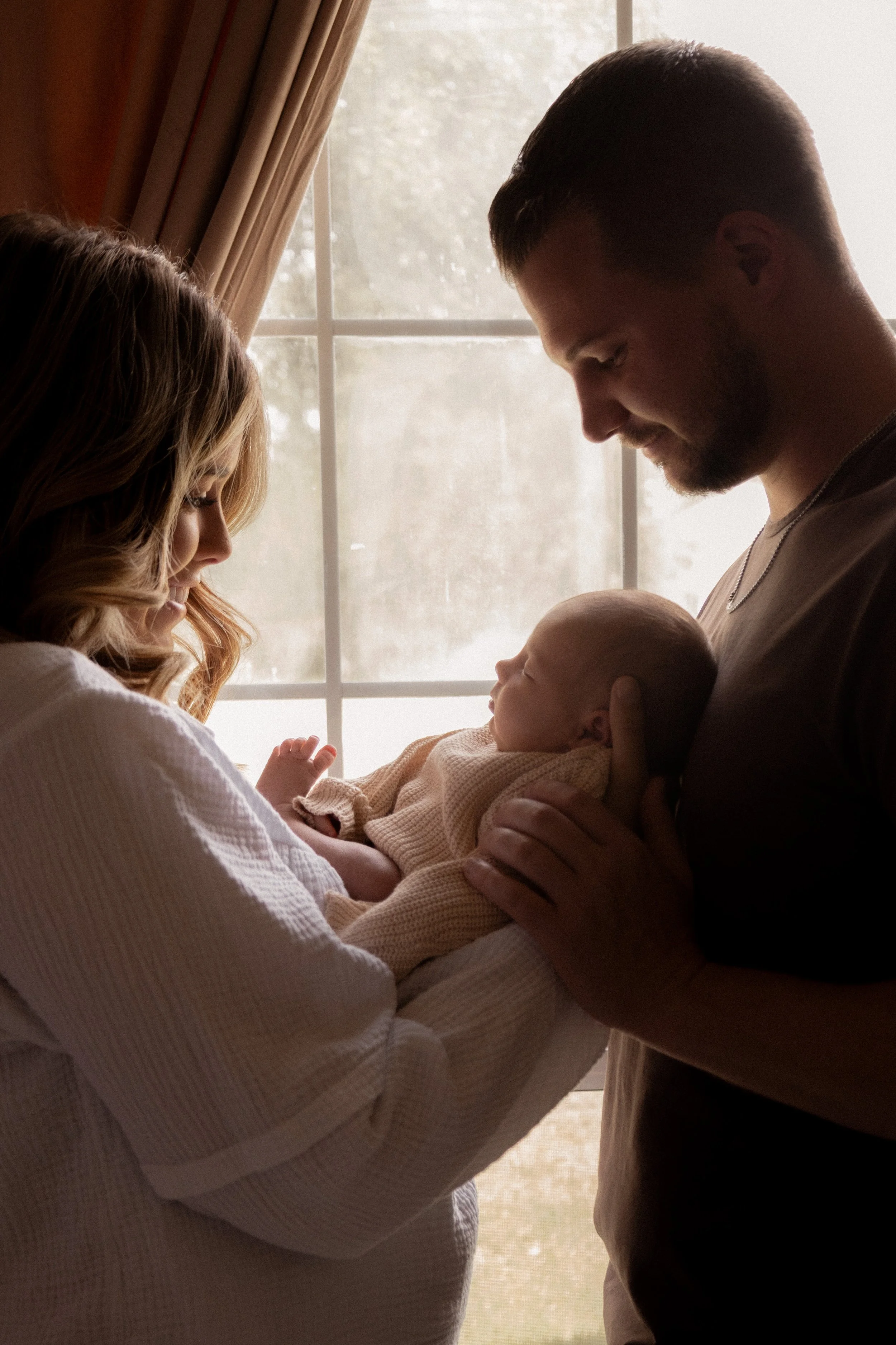 A woman, a man, and a baby are standing by a window with daylight coming through. The woman is holding the baby, and the man is gently touching the baby's head. They are looking at the baby and smiling softly.
