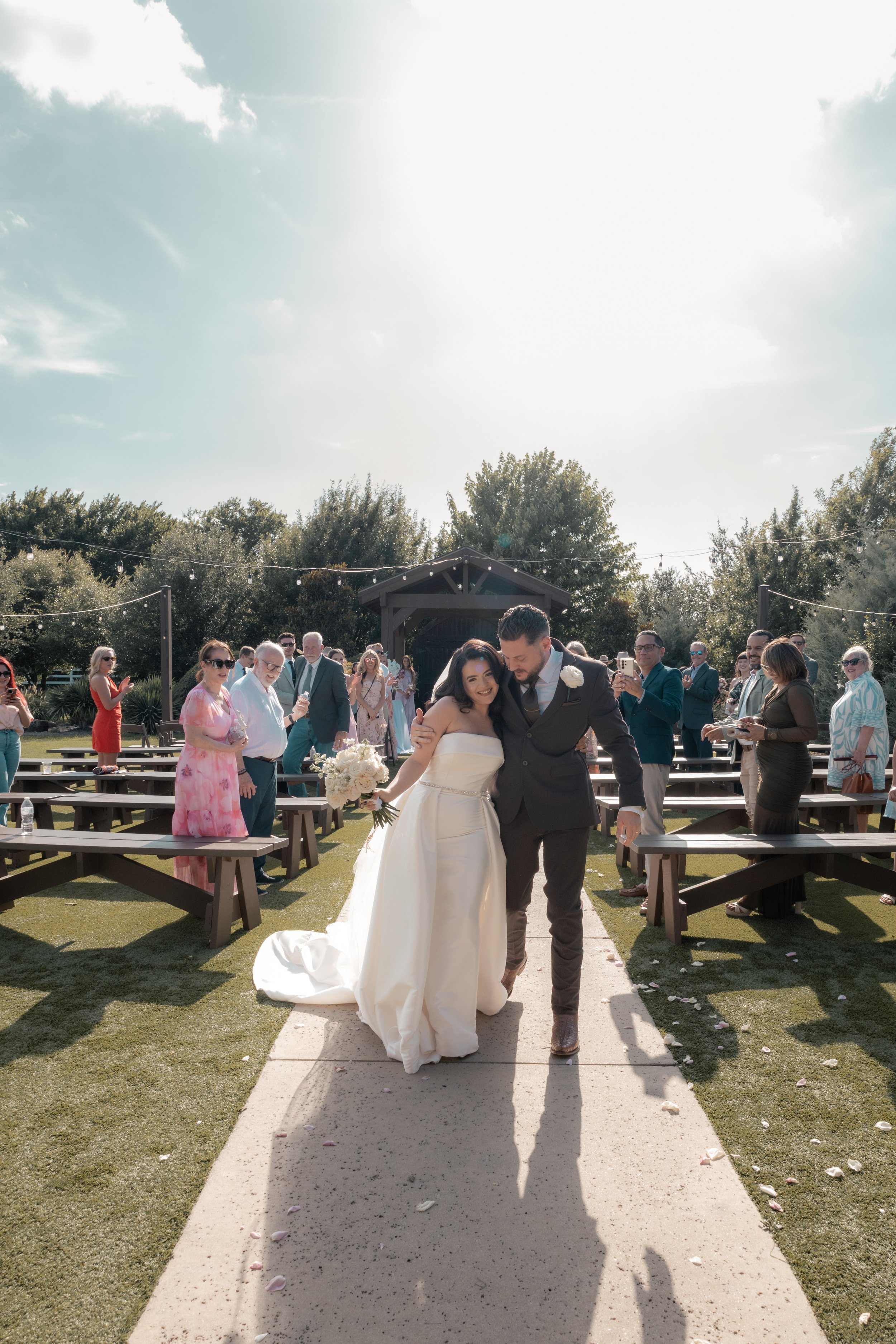 A newlywed couple is walking down an aisle outdoors, smiling and embracing. The bride is in a white wedding gown holding a bouquet, and the groom is in a dark suit. Guests are standing and clapping on either side, with some taking photos. The ceremony appears to be taking place in a garden with trees, a small pavilion, and string lights in the background. The sky is partly cloudy and bright.