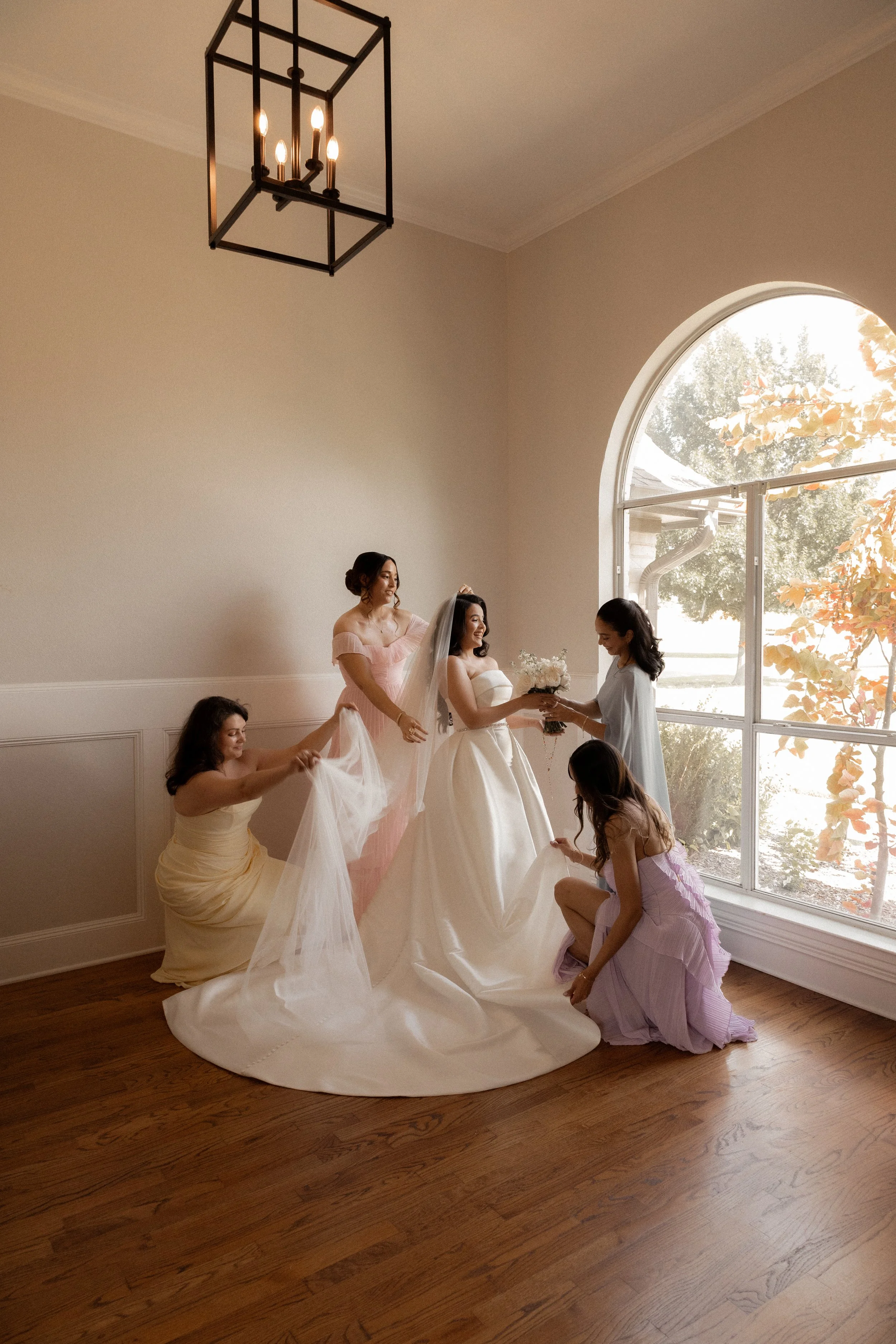 A group of women, including a bride in a white wedding dress, and bridesmaids in pastel dresses, preparing for a wedding ceremony inside a bright room with a large arched window and a hanging geometric chandelier.