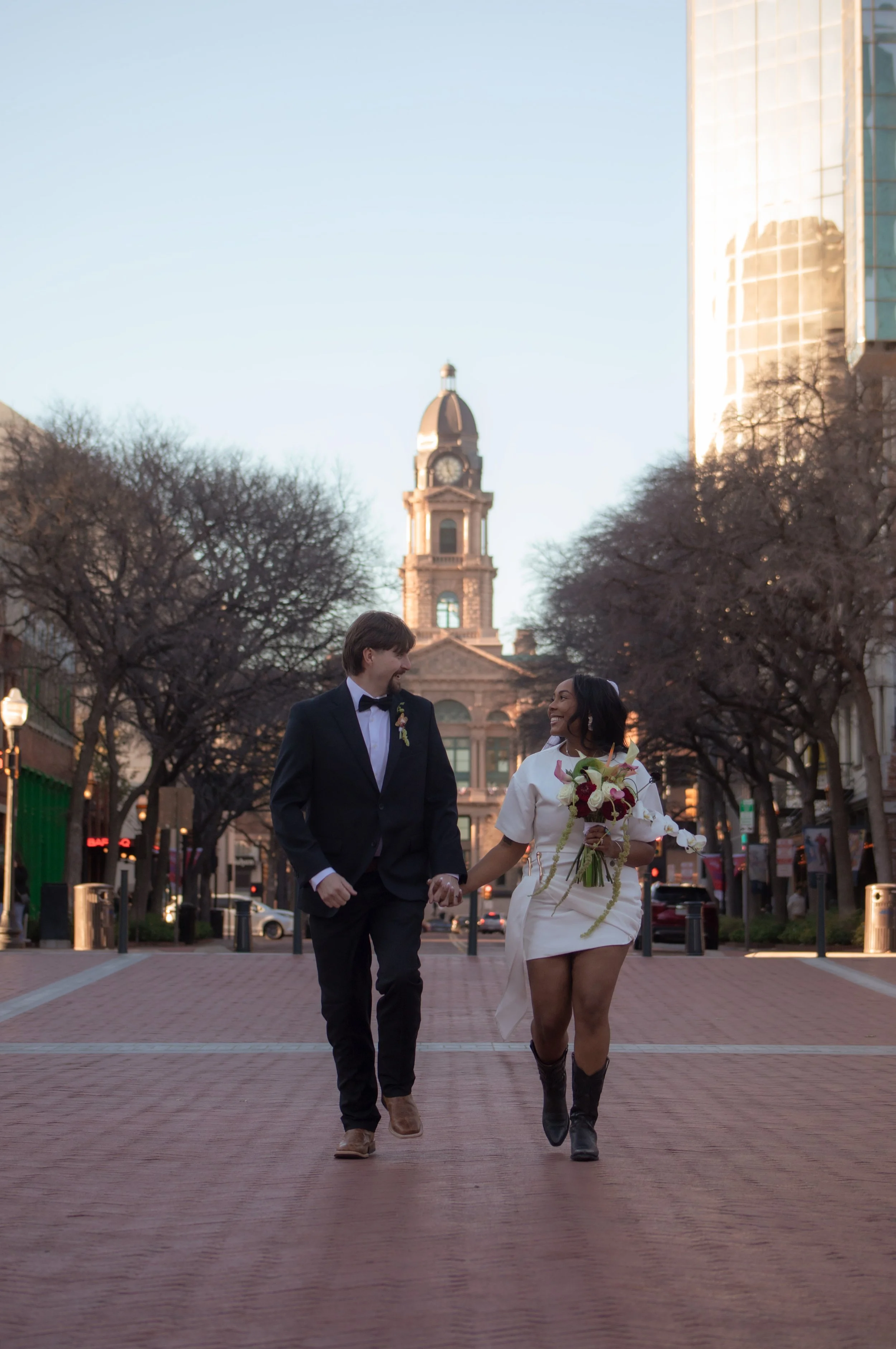 A newly married couple walking hand in hand on a city street at sunset, with a historic building with a clock tower in the background.