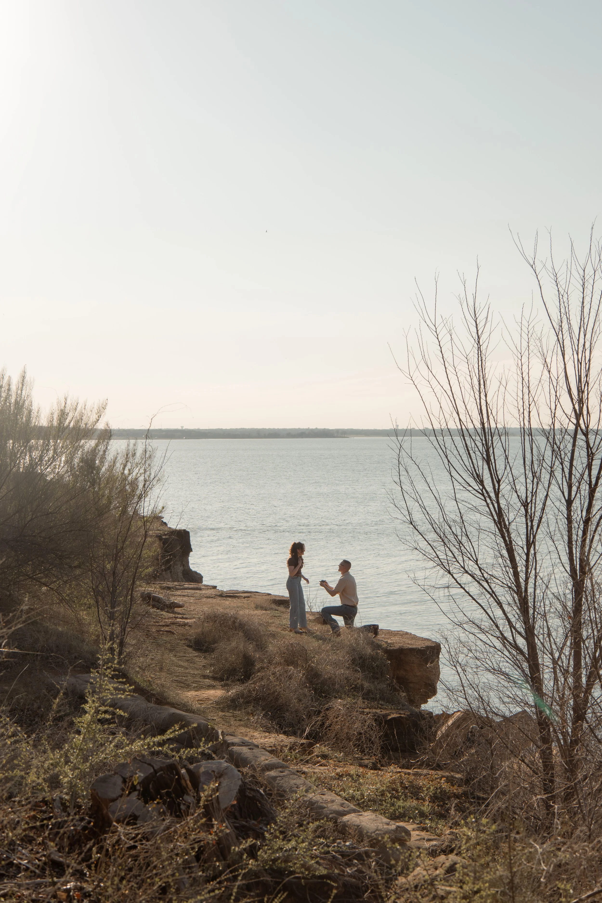 A man on one knee proposing to a woman on a rocky cliff near a body of water, with leafless trees and dry vegetation in the surrounding landscape.