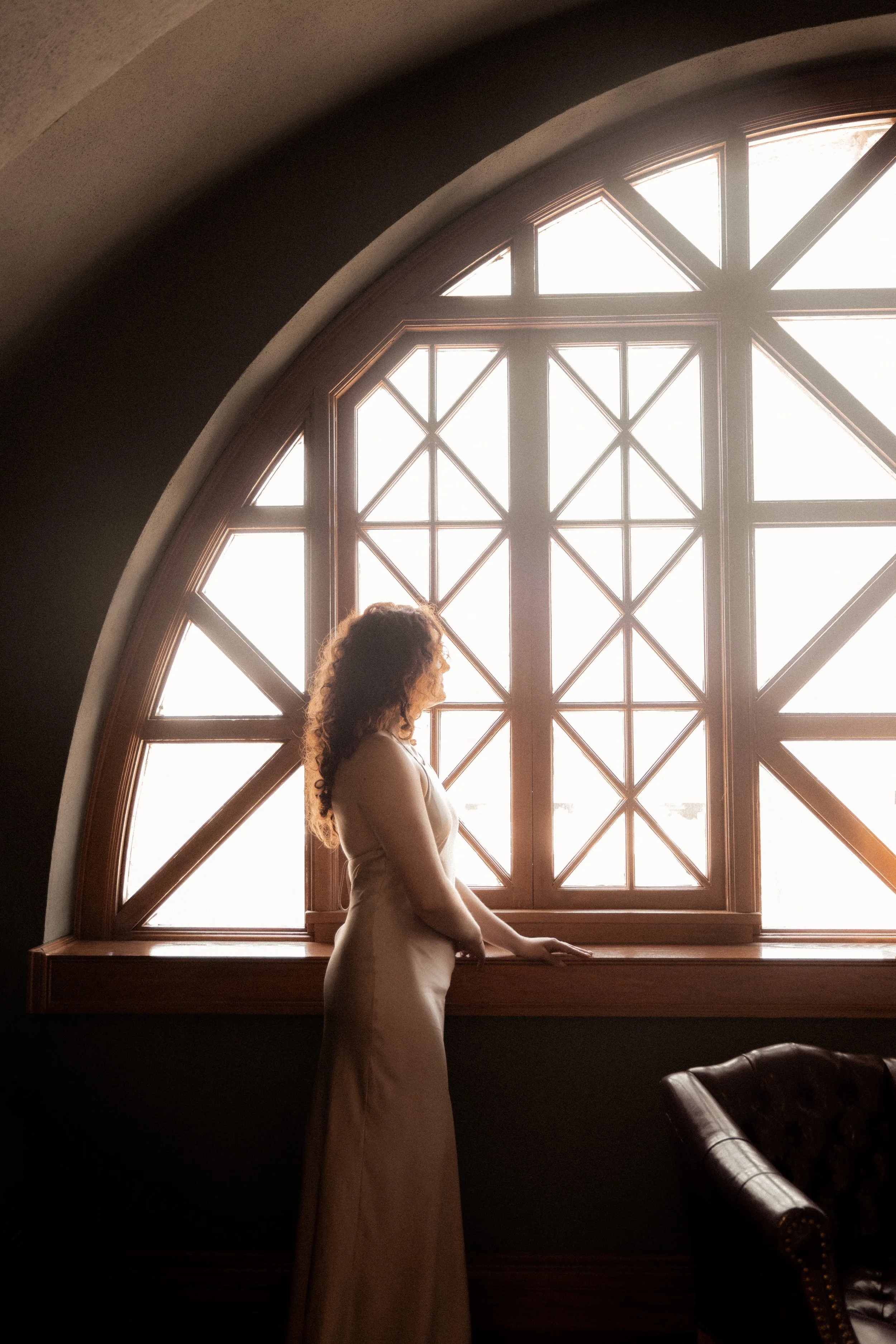 A woman with curly hair in a long dress standing in profile near a large, arched window with wooden framing, with sunlight streaming in.