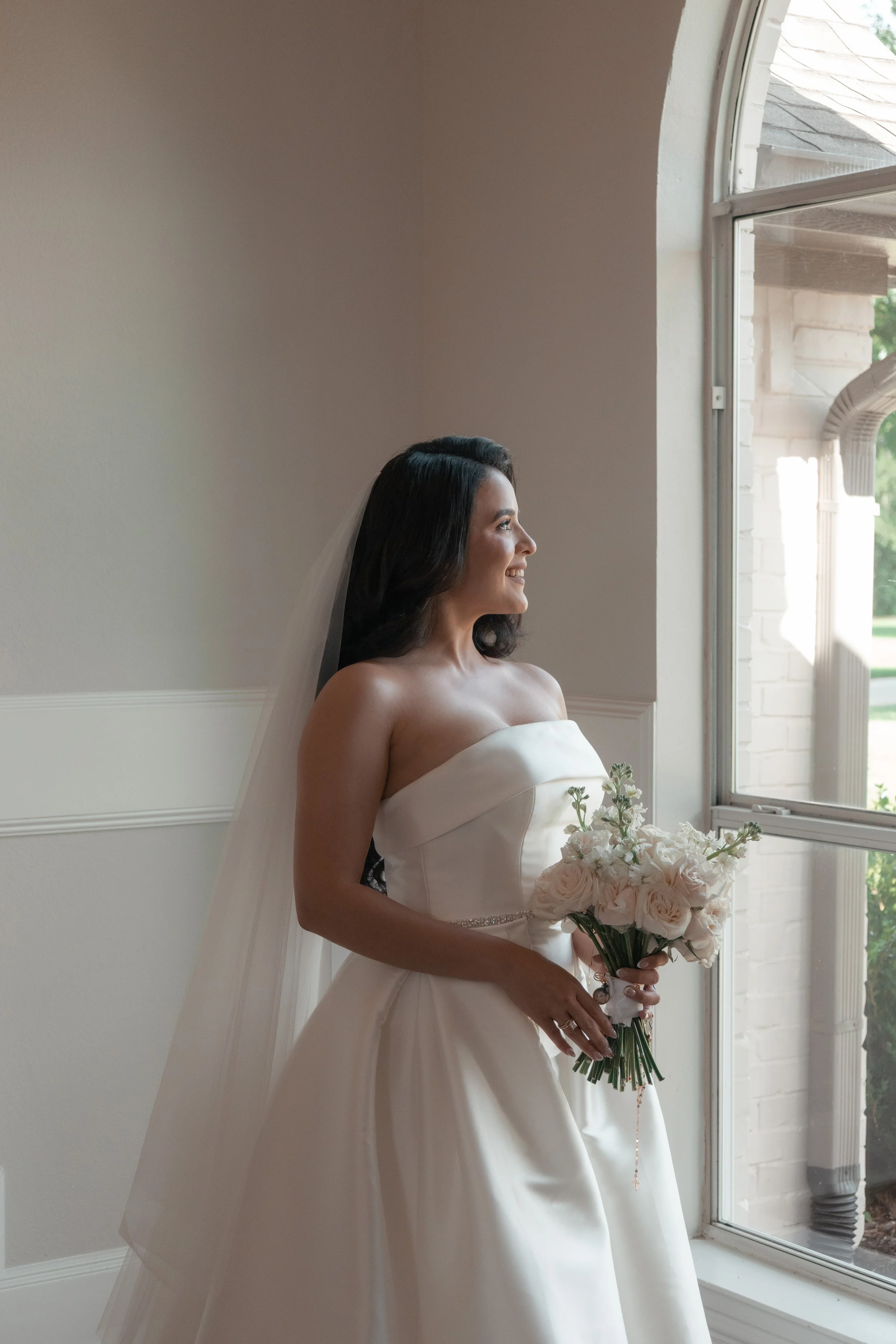 A bride with dark, wavy hair, wearing a white strapless wedding dress, holding a bouquet of pale pink roses, standing near an open window, smiling as she looks outside.