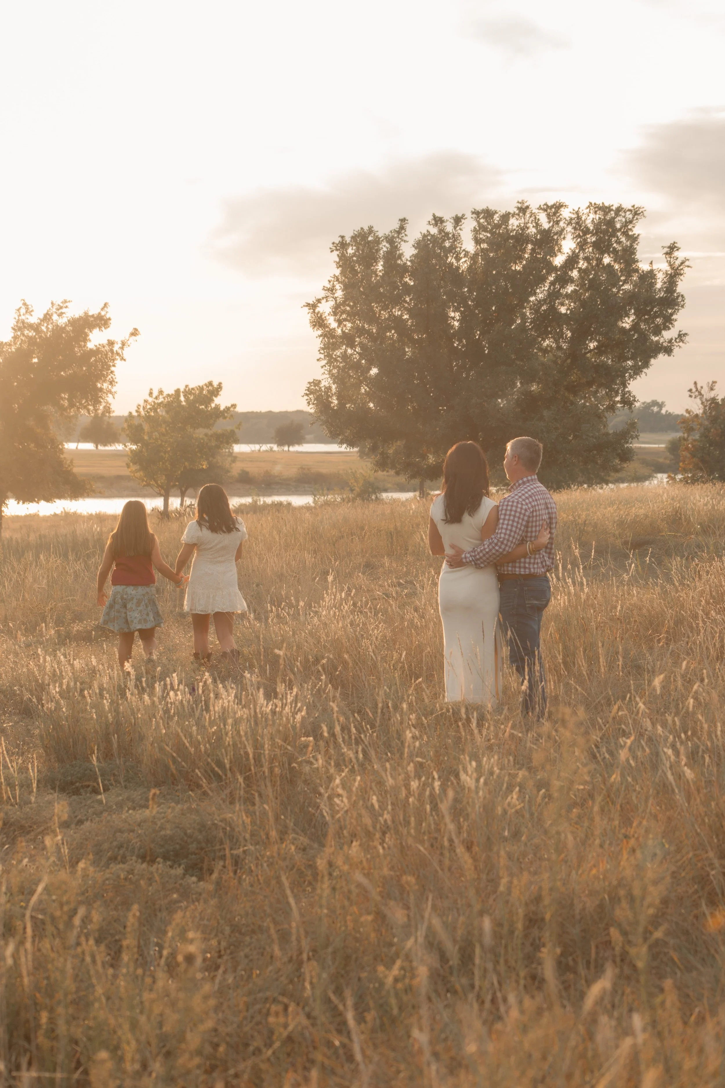 A group of four people in a field during sunset, with large trees and water in the background. Two women in the foreground, one holding the other, with two women walking away in the distance.