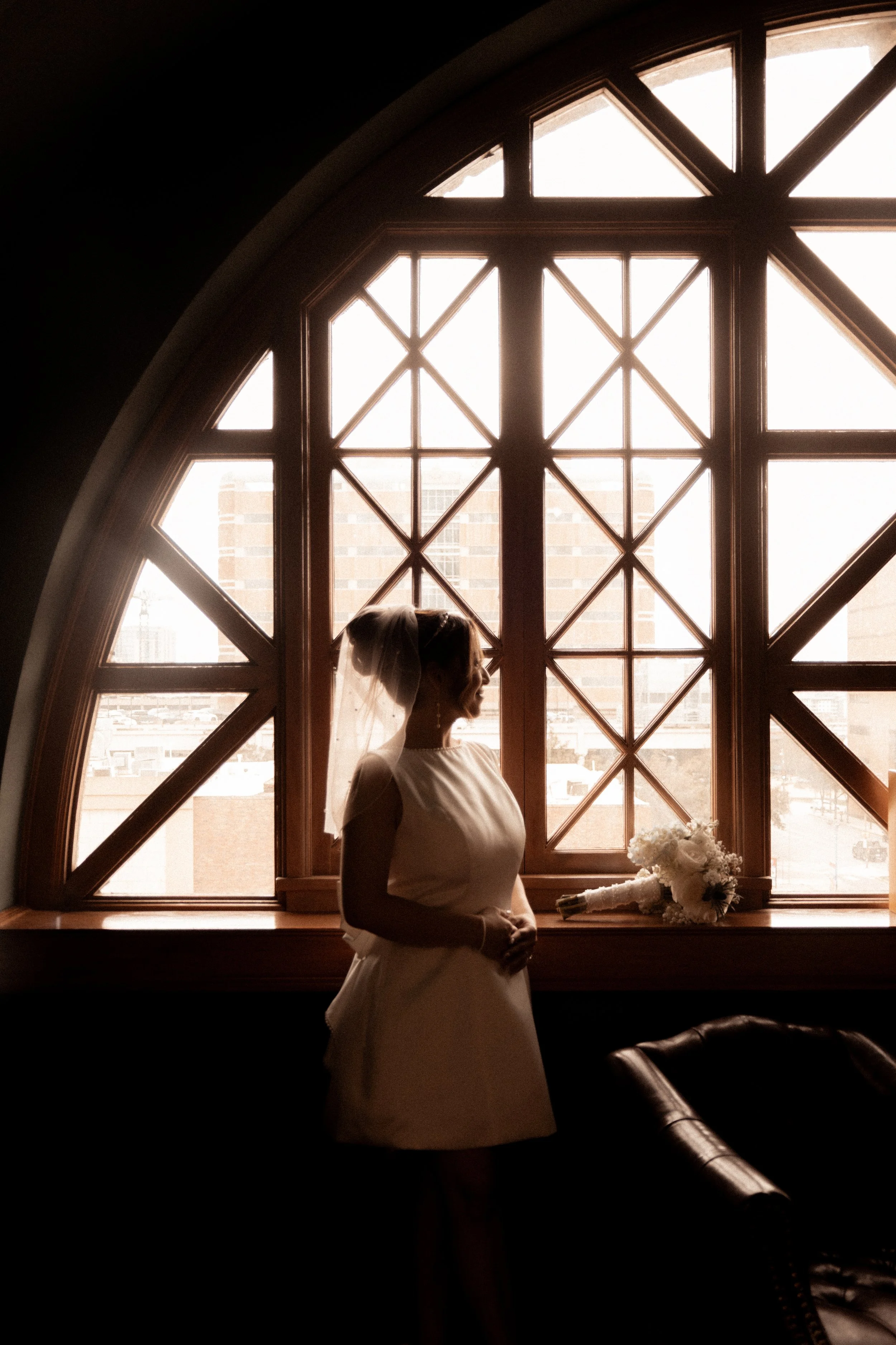 A bride standing indoors near a large arched window, holding a bouquet, with sunlight shining through the window, creating a silhouette effect.
