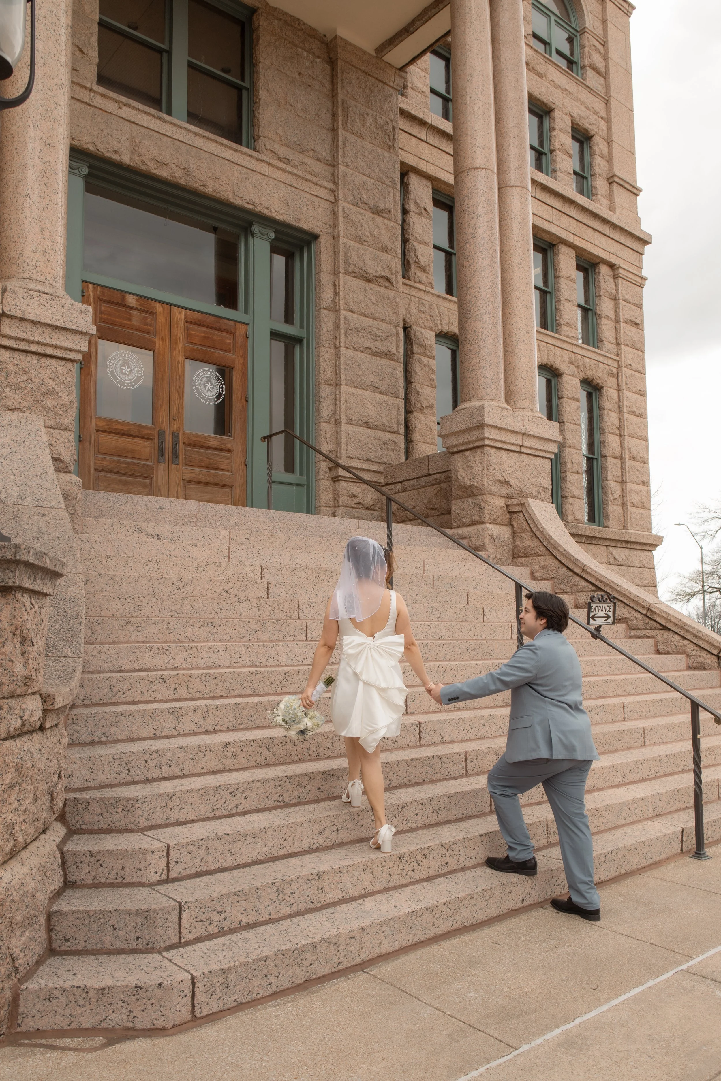 A bride wearing a white dress and veil is ascending outdoor stone steps, holding a bouquet, as a groom in a gray suit kneels and holds her hand, in front of a large historic building with brown double doors.