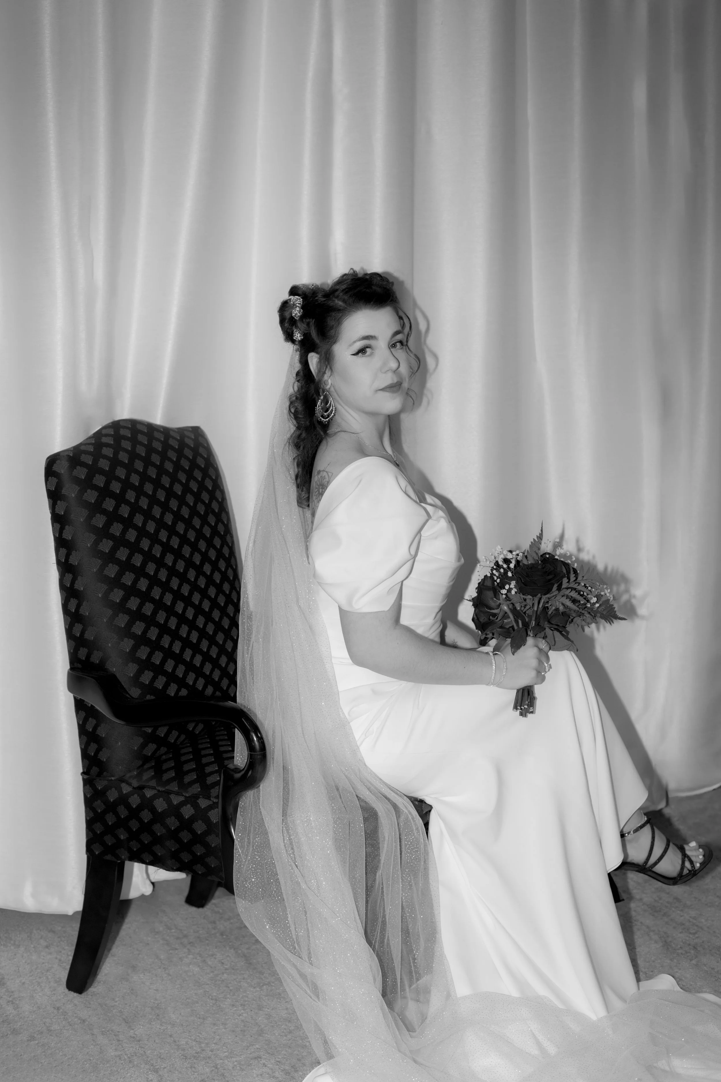 Black and white photo of a woman in a wedding dress sitting on a chair, holding a bouquet, with a curtain backdrop.