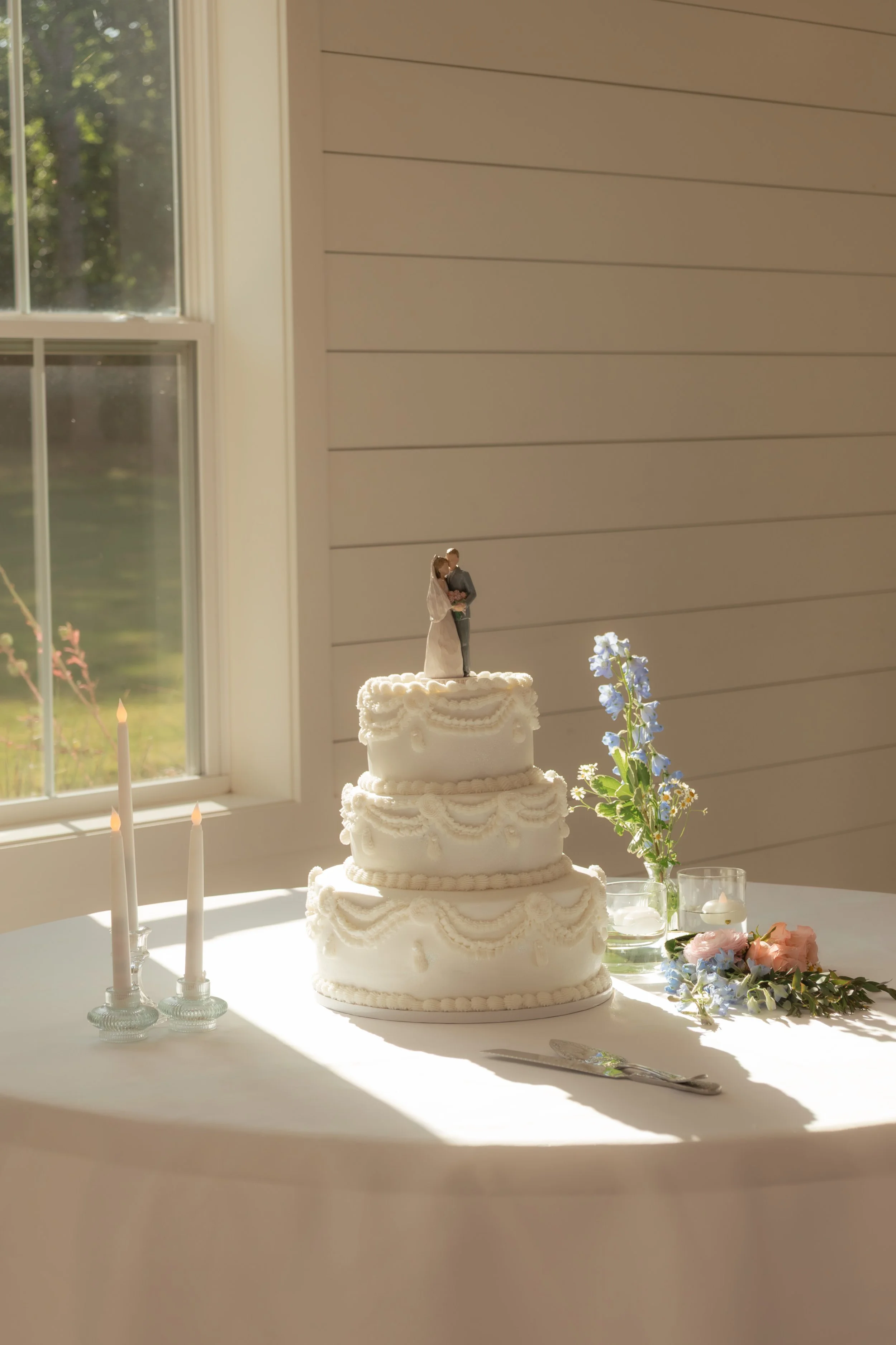 Wedding cake with three layers decorated with white icing rosettes and piping, topped with a figurine of a bride and groom, located on a white table near a window with sunlight, alongside a vase of blue and pink flowers and candles.