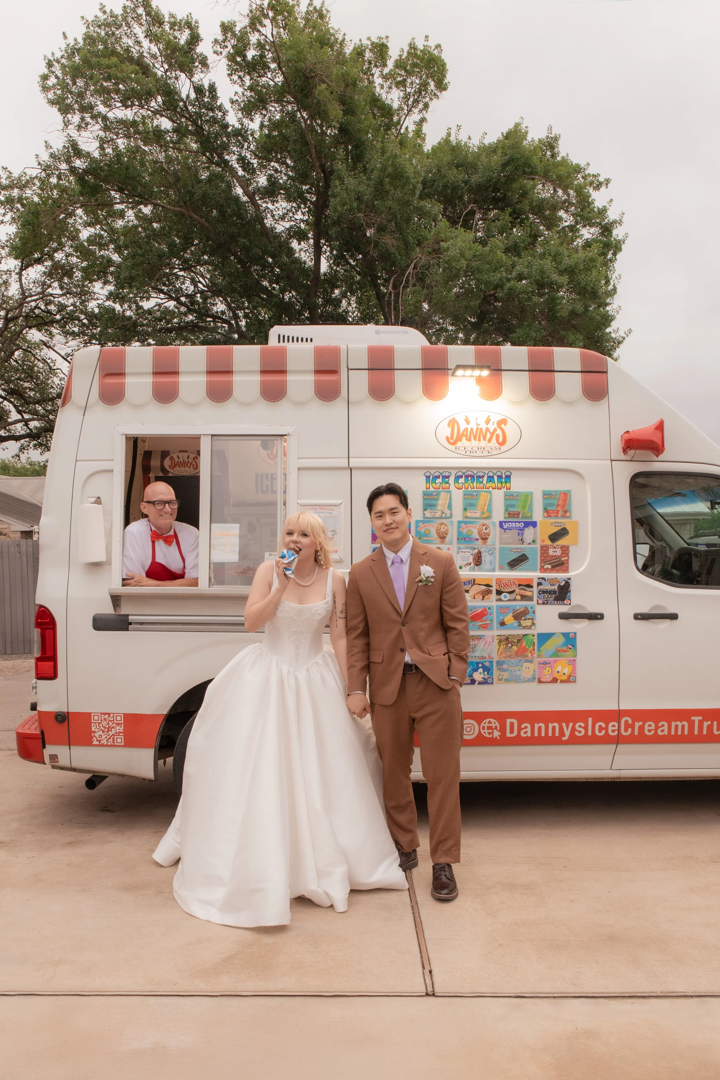 A bride and groom holding hands in front of an ice cream truck, with a woman eating ice cream and an ice cream vendor smiling inside the truck. There are large trees in the background and a cloudy sky.