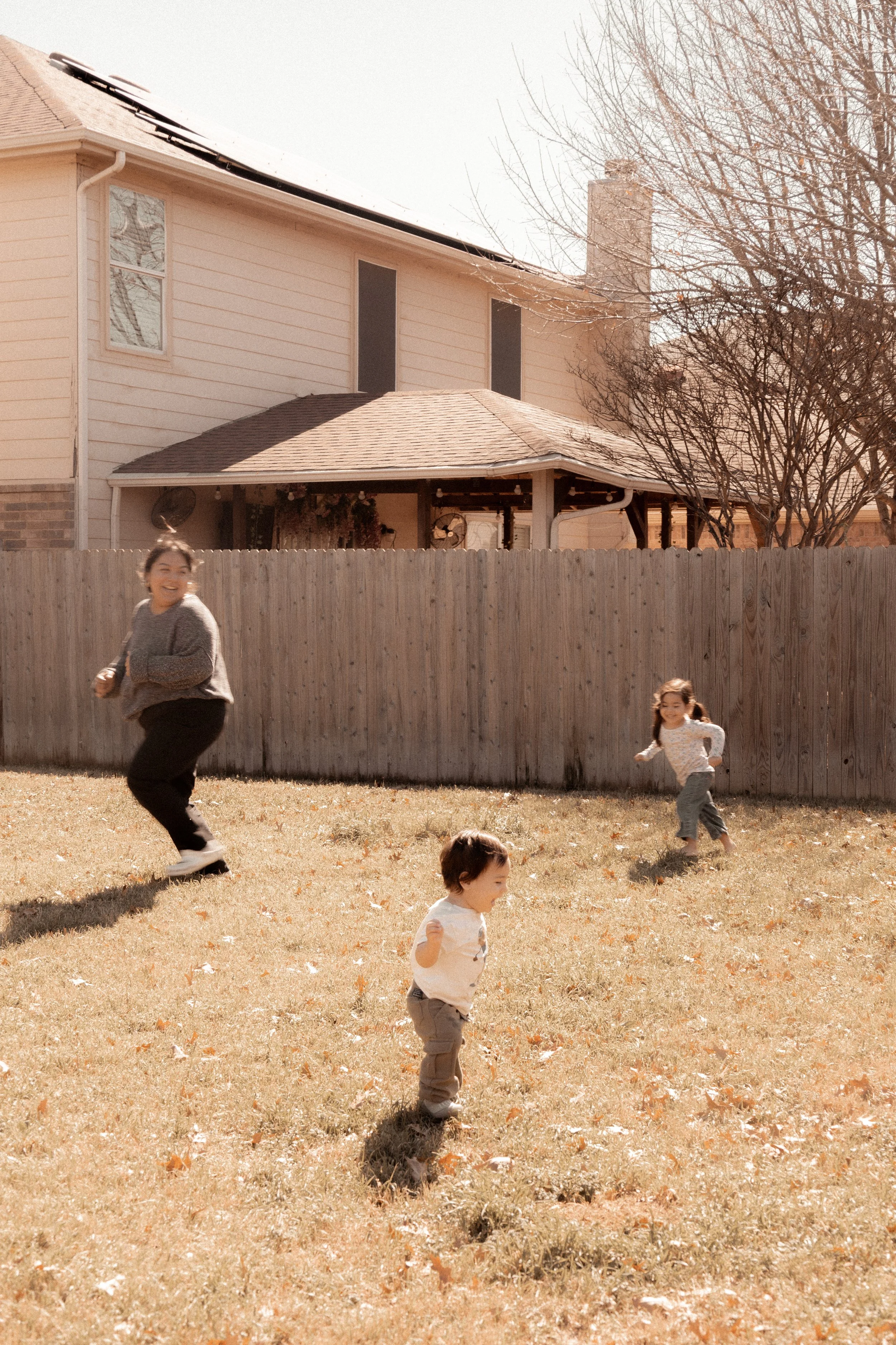 A woman and two children playing and running on a grassy backyard with a wooden fence and a house in the background under a clear sky.