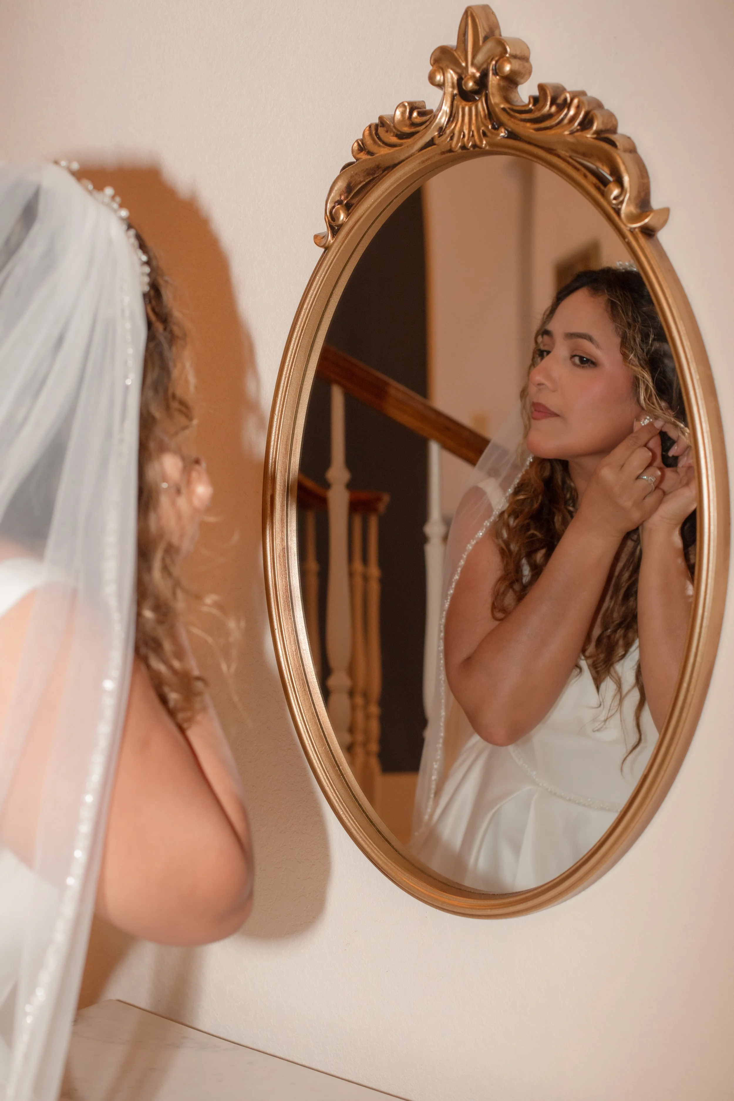 A bride putting on earrings while looking in an oval mirror with a decorative gold frame.