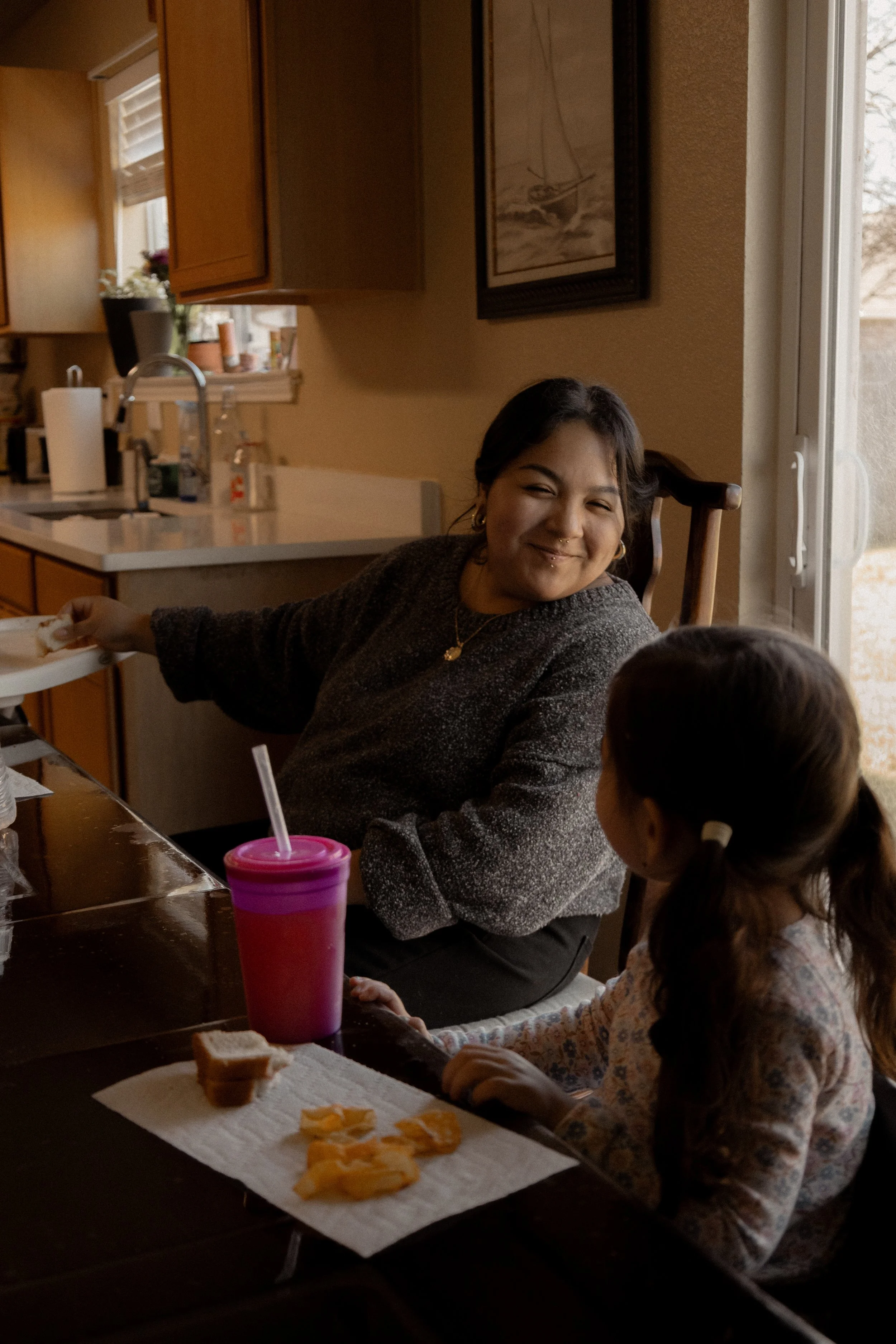 A woman and a young girl having a conversation at a kitchen table, with snacks, a pink cup, and a slice of bread on a paper towel.