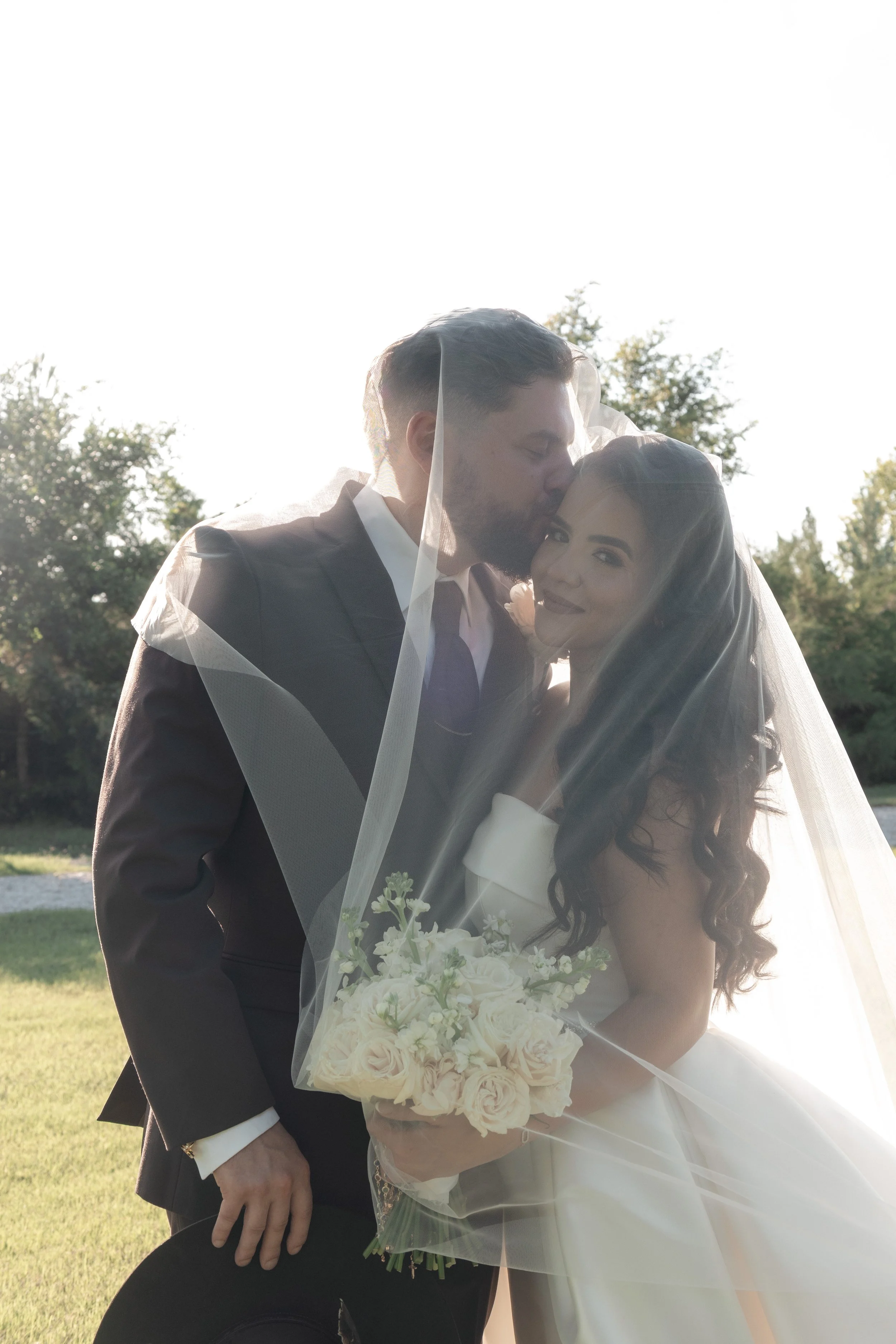 A newlywed couple kissing outdoors, with the groom in a suit and the bride holding a bouquet of white roses, all under bright sunlight with trees in the background.