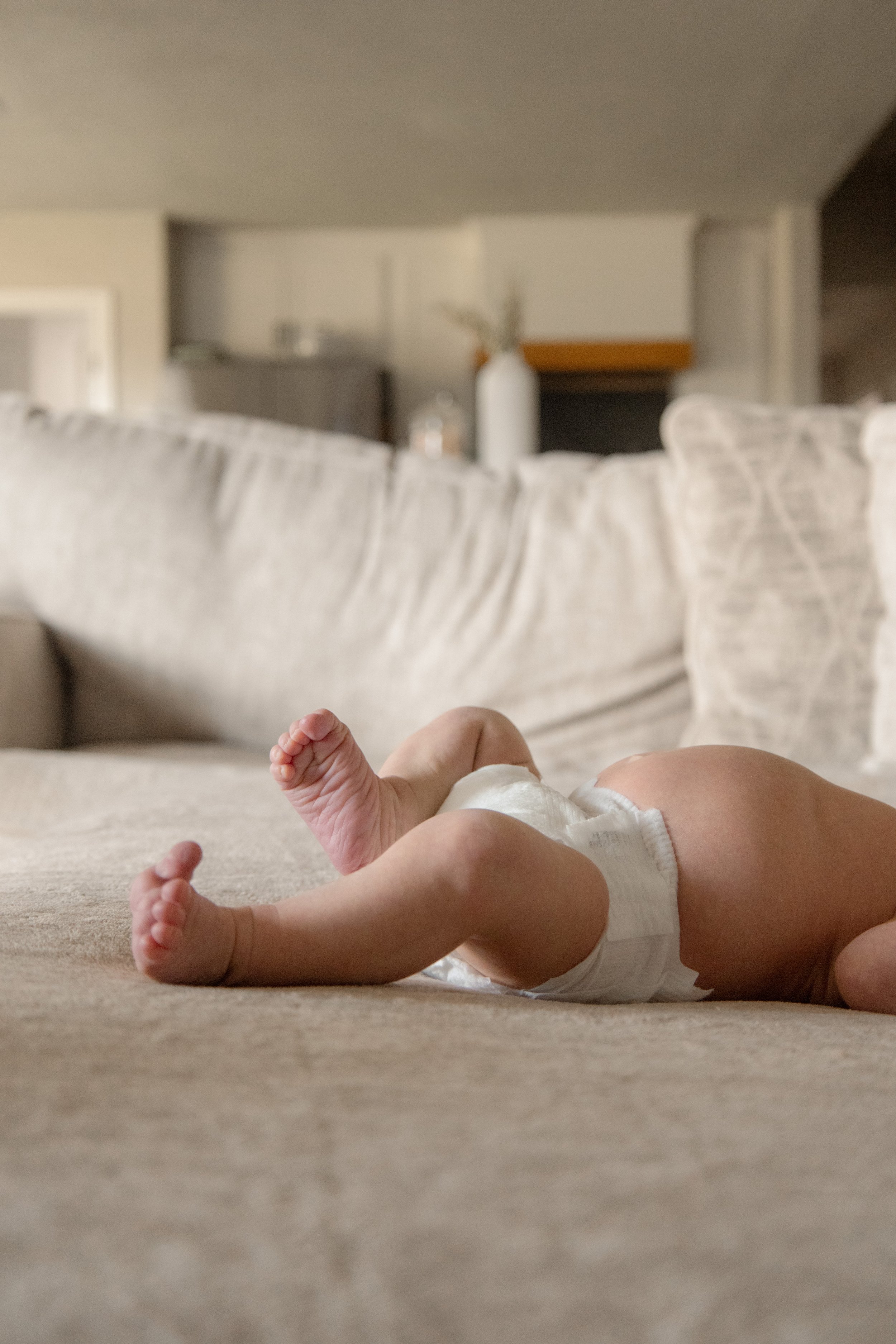 Close-up of a baby lying on a beige carpet, with a beige sofa and a blurred background of a living room.