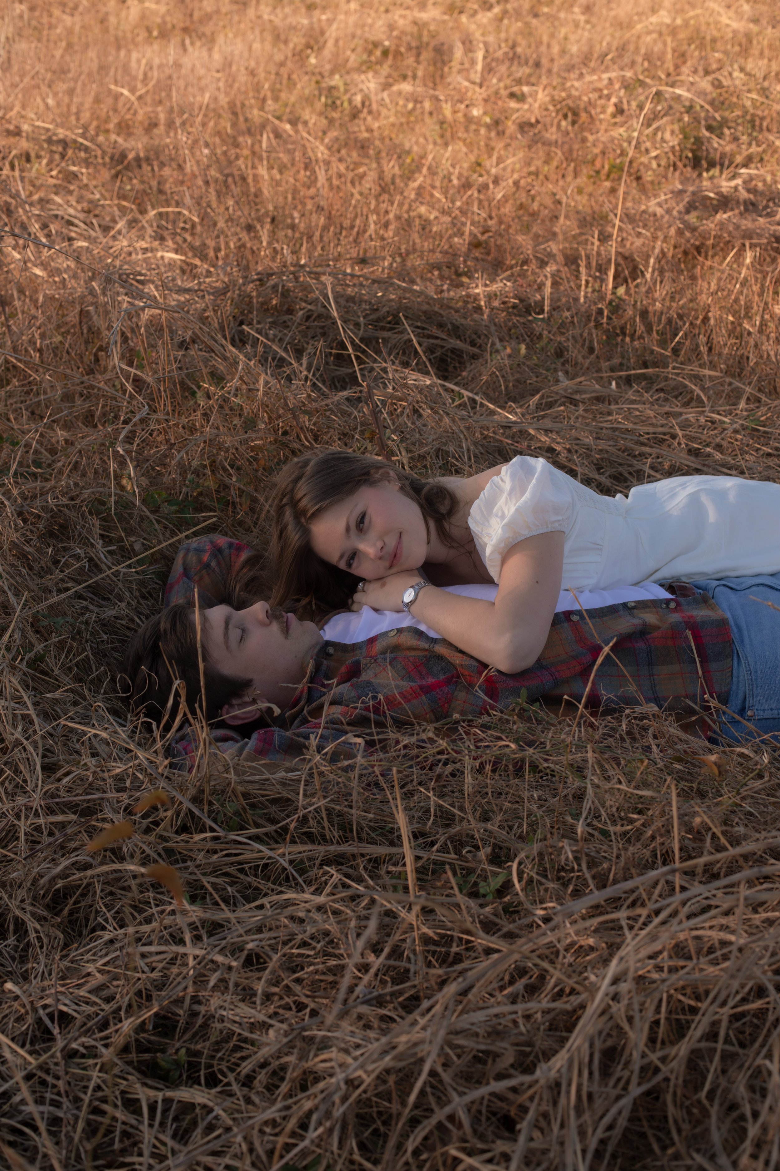 A couple laying on the ground in a field of dry grass, with the woman resting her head on the man's chest and looking at him.