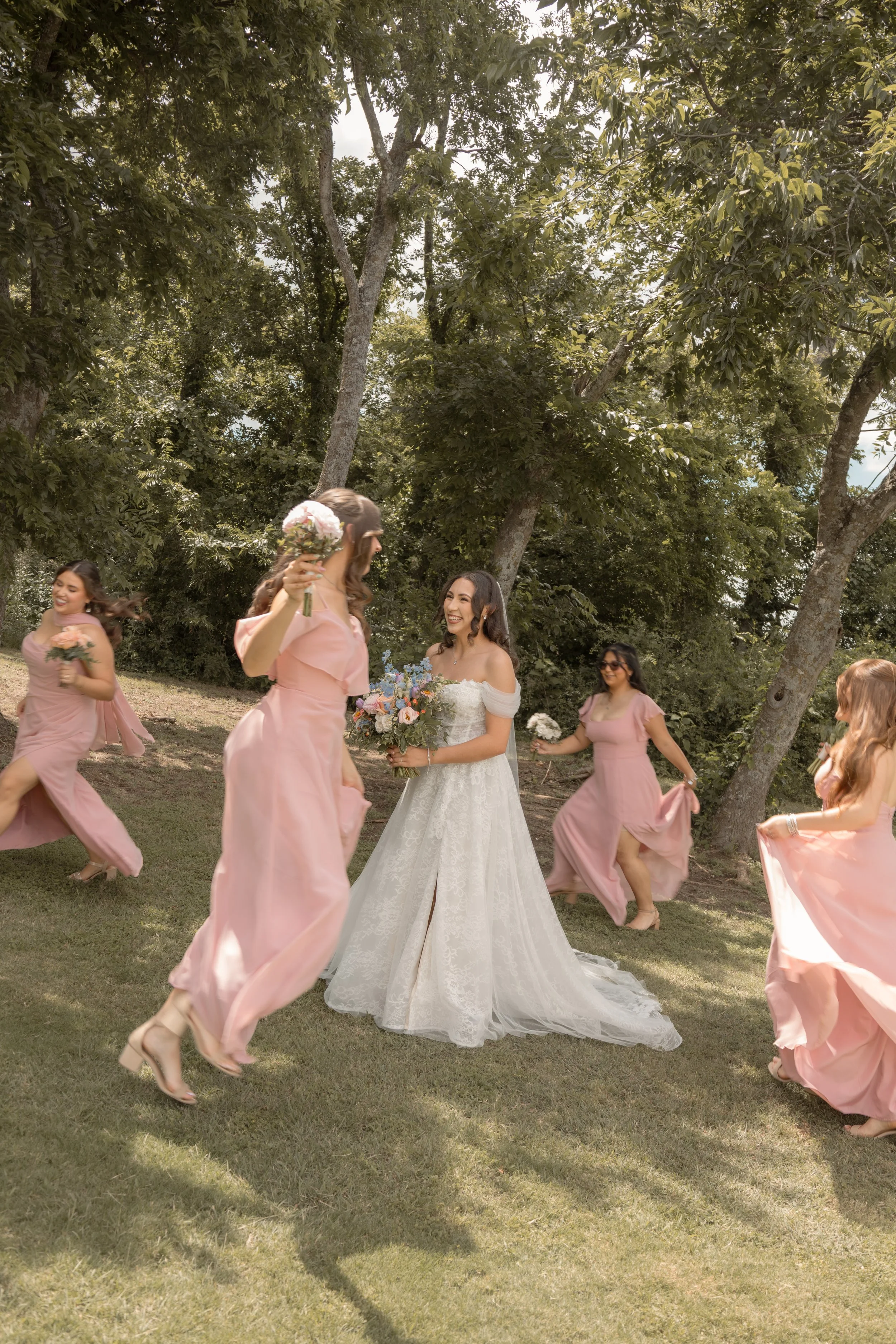A bride in a white wedding gown holding a bouquet surrounded by bridesmaids in pink dresses dancing outdoors under trees.