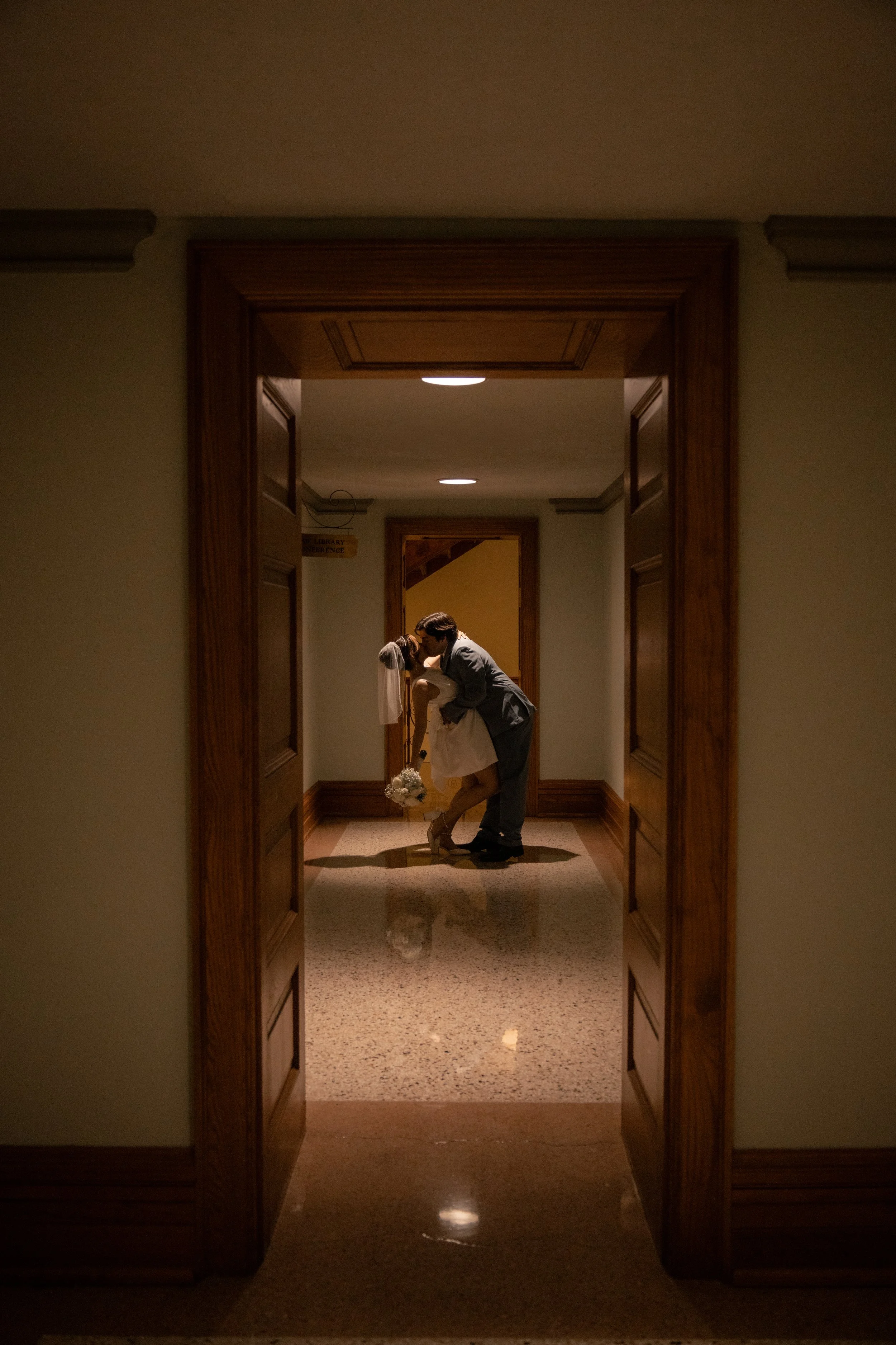 A bride and groom sharing a kiss in a dimly lit hallway, framed by open double doors, with a woman holding a bouquet nearby.