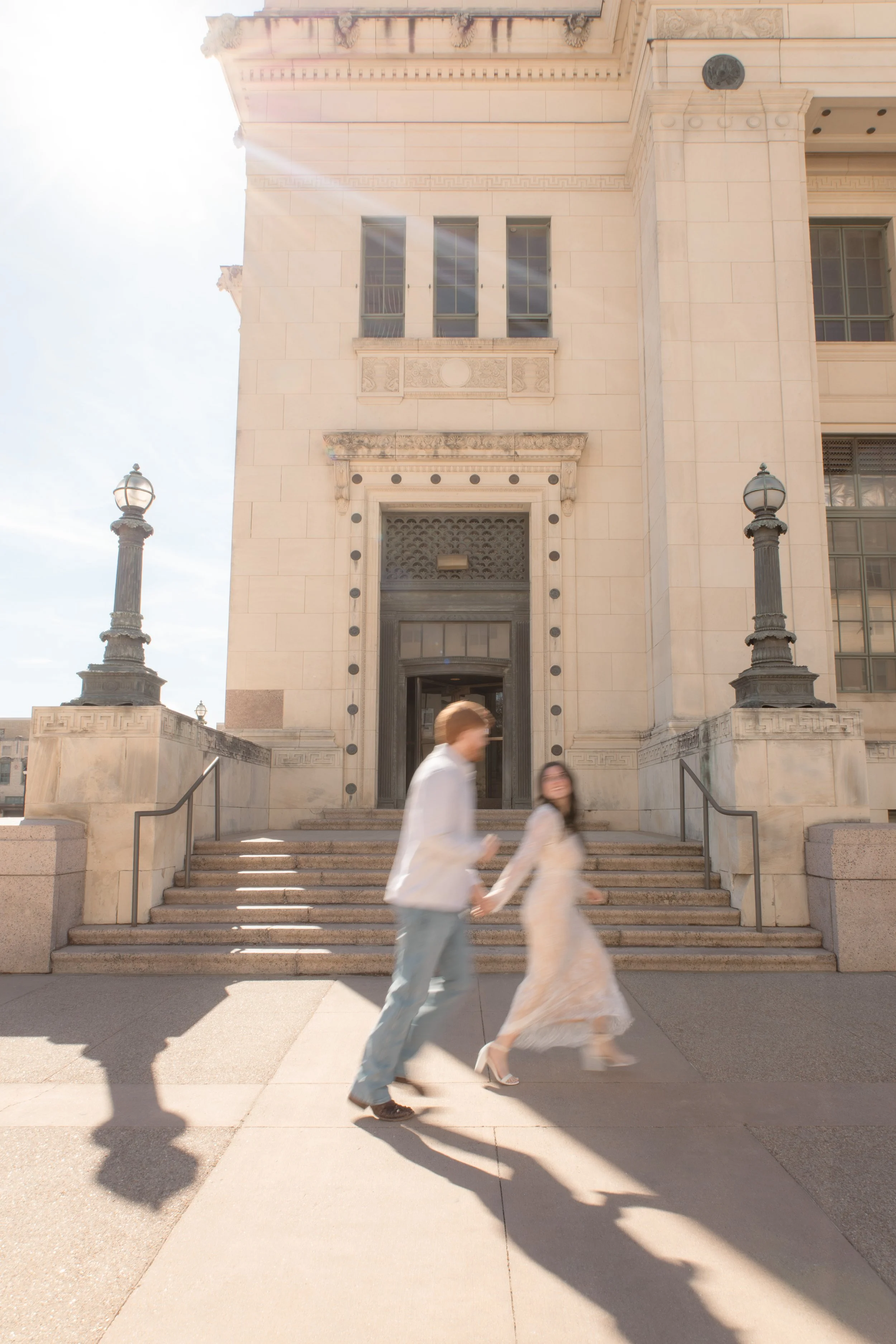 A couple is walking hand-in-hand in front of a historic building with steps and ornate lamps, sunlight shining from the upper left corner.