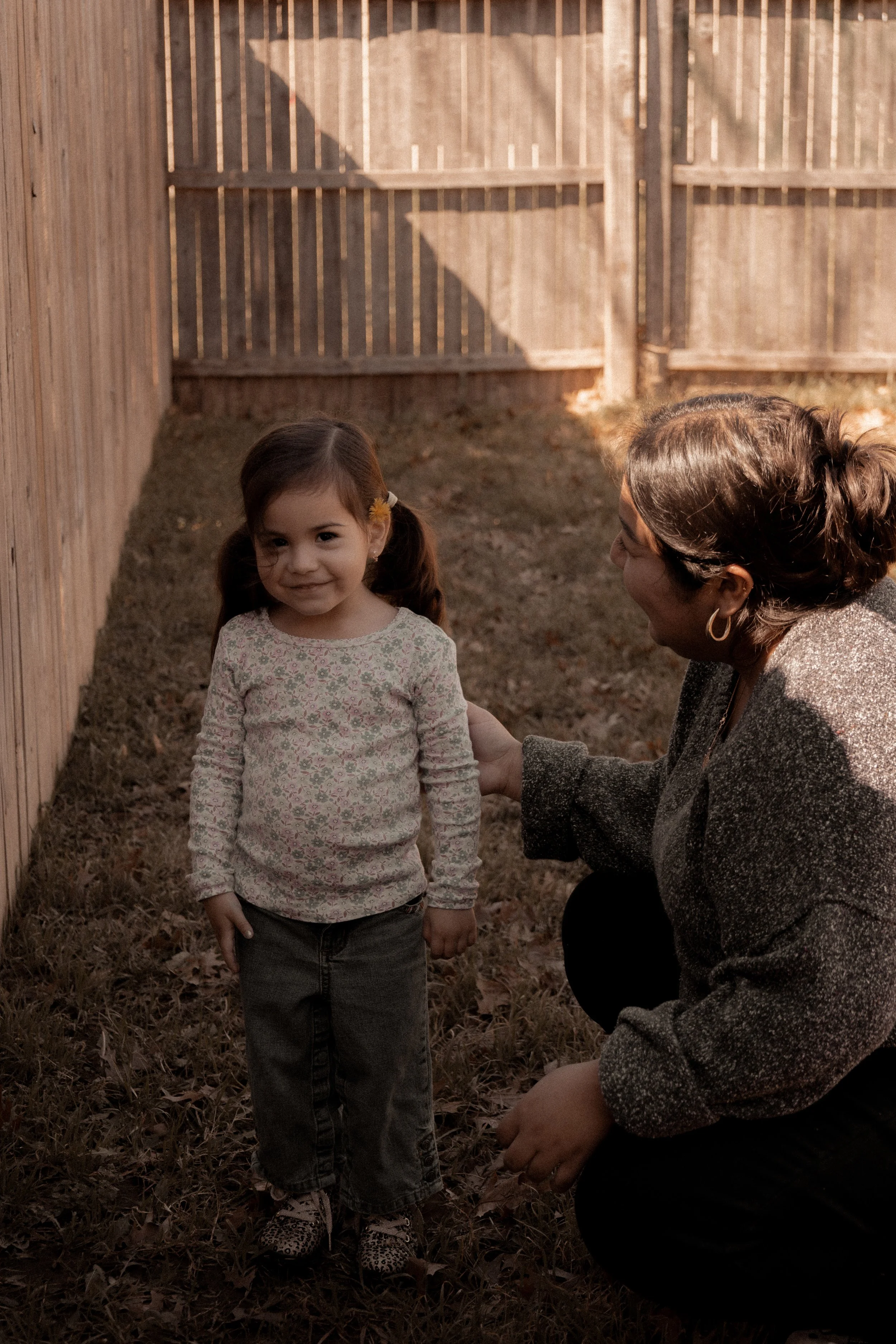 A young girl with pigtails and a floral sweater standing outdoors next to an adult woman who is crouching and smiling at her. They are near a wooden fence with a patch of grass and fallen leaves on the ground.