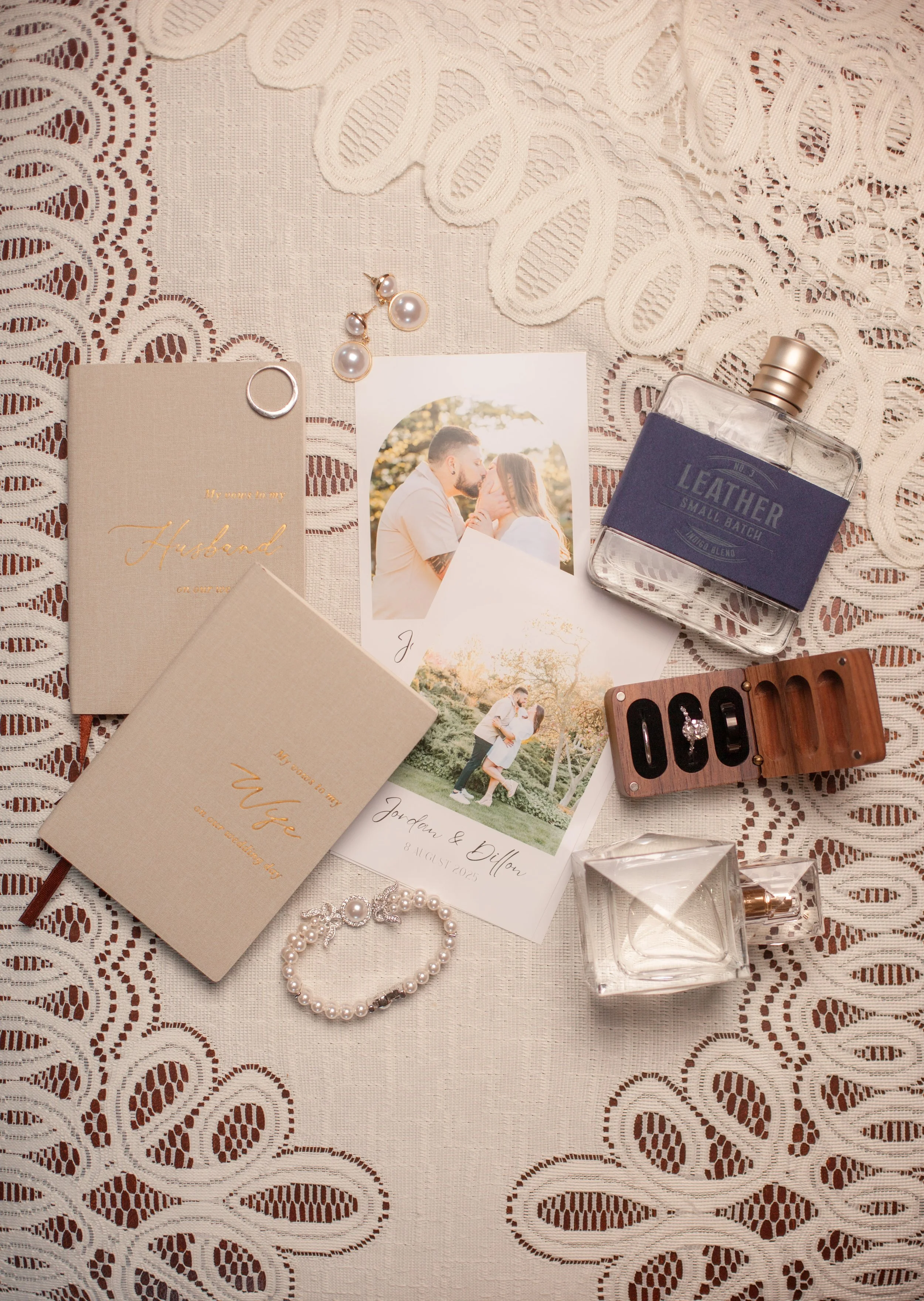 Wedding or engagement invitation cards, photographs of a couple, jewelry including pearl earrings and a pearl bracelet, a perfume bottle, a leather notebook, a ring box with an engagement ring, a glass perfume bottle, and a small glass container, all