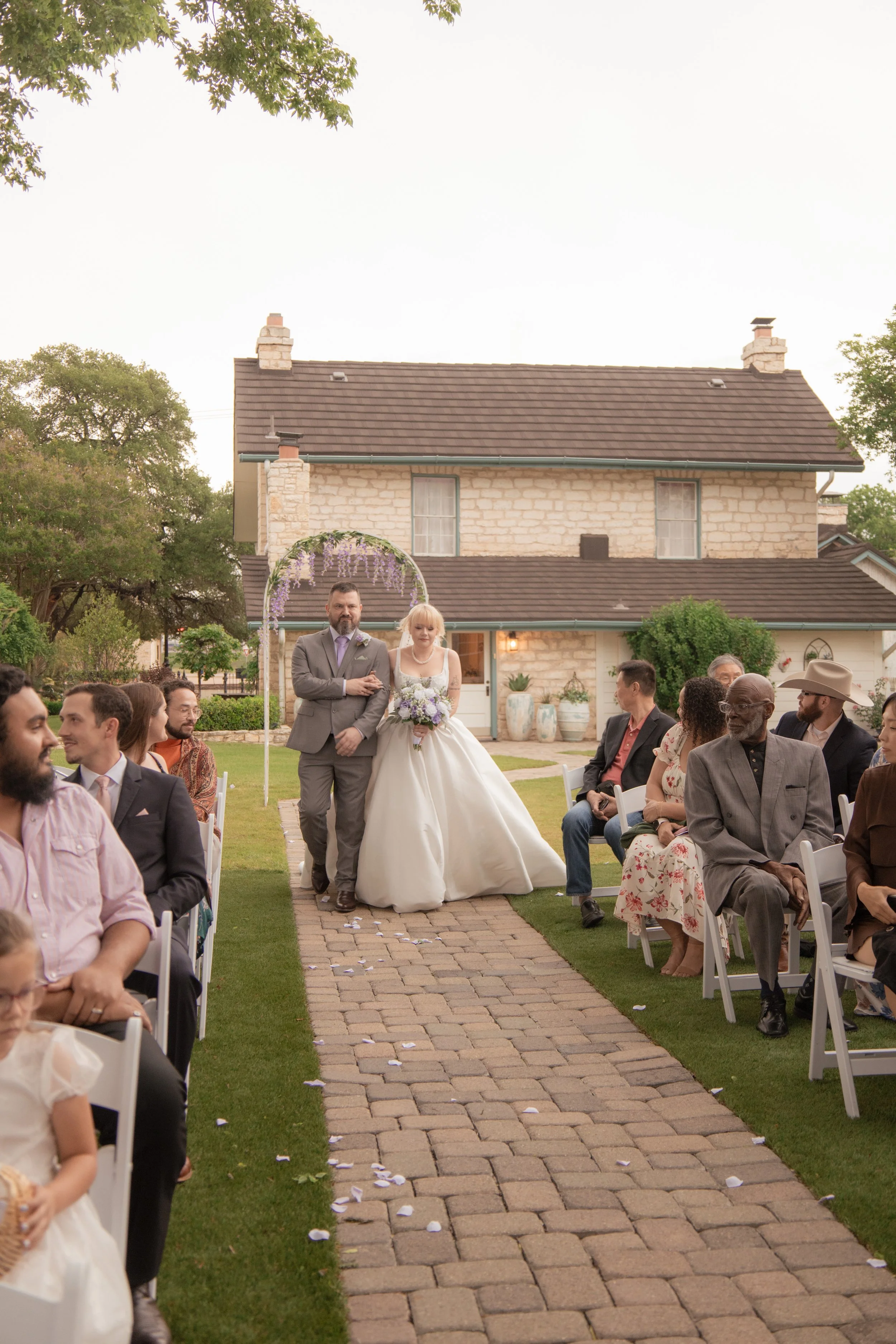 Wedding ceremony outdoors with a bride and a man walking down the aisle. Guests seated on either side and a house with a garden in the background.