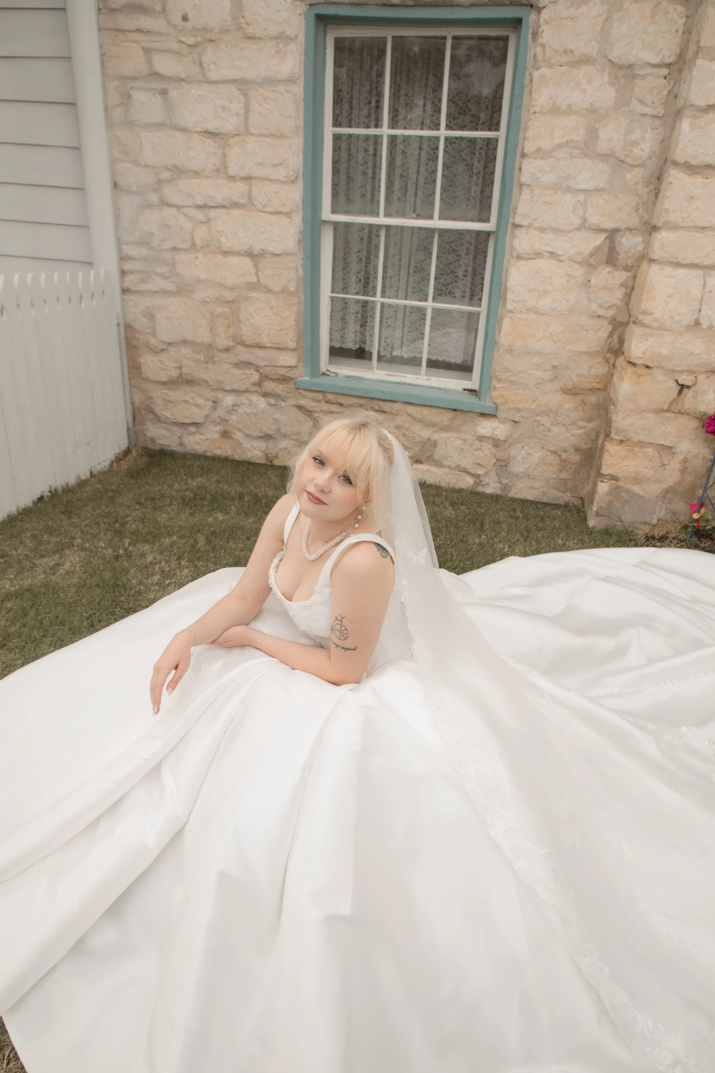 A woman in a wedding dress sitting on a white wedding gown outdoors in front of a stone wall and window.