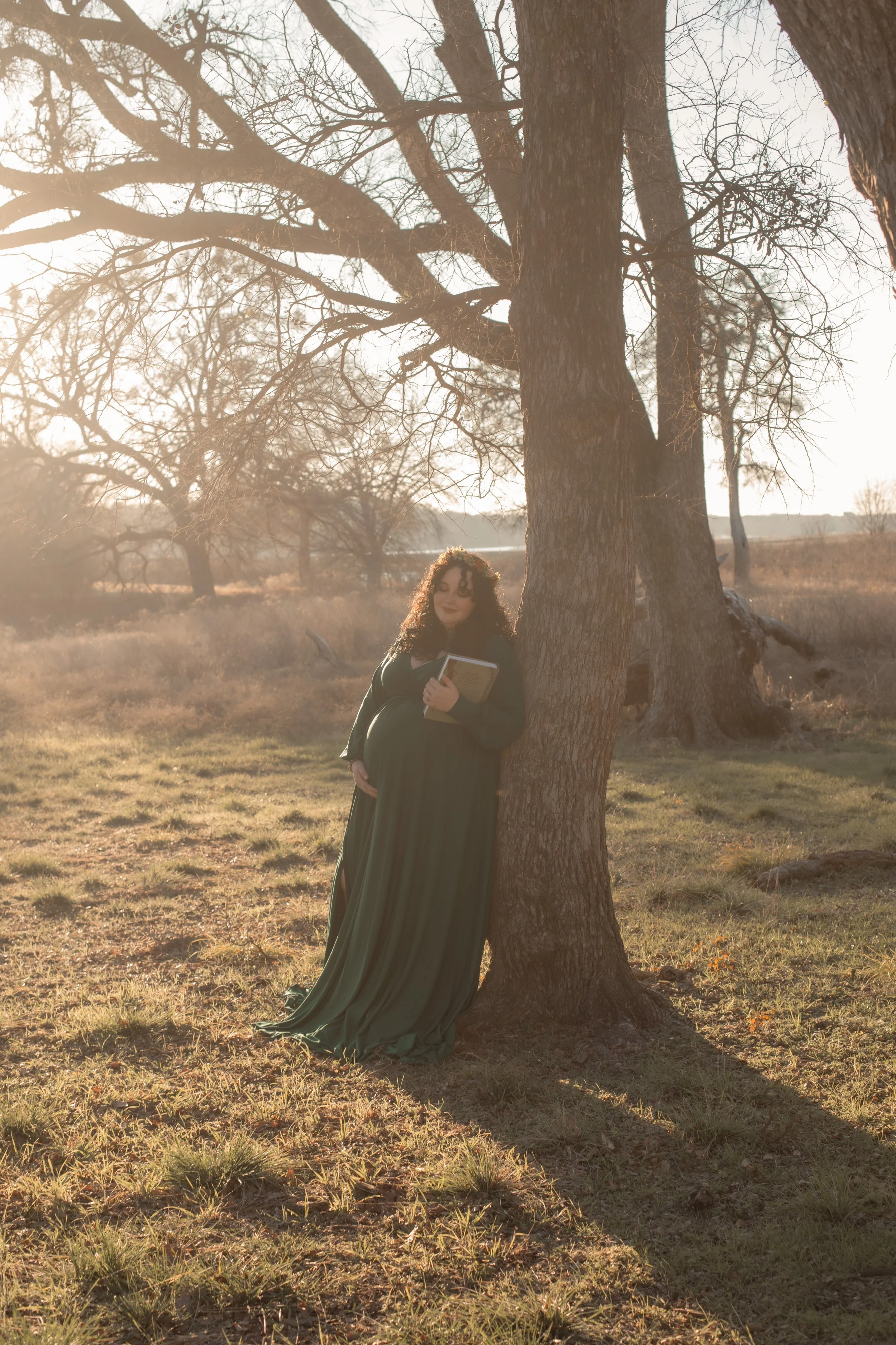 A pregnant woman in a long green dress stands beside a large tree in a field during sunset, holding a book and smiling softly.