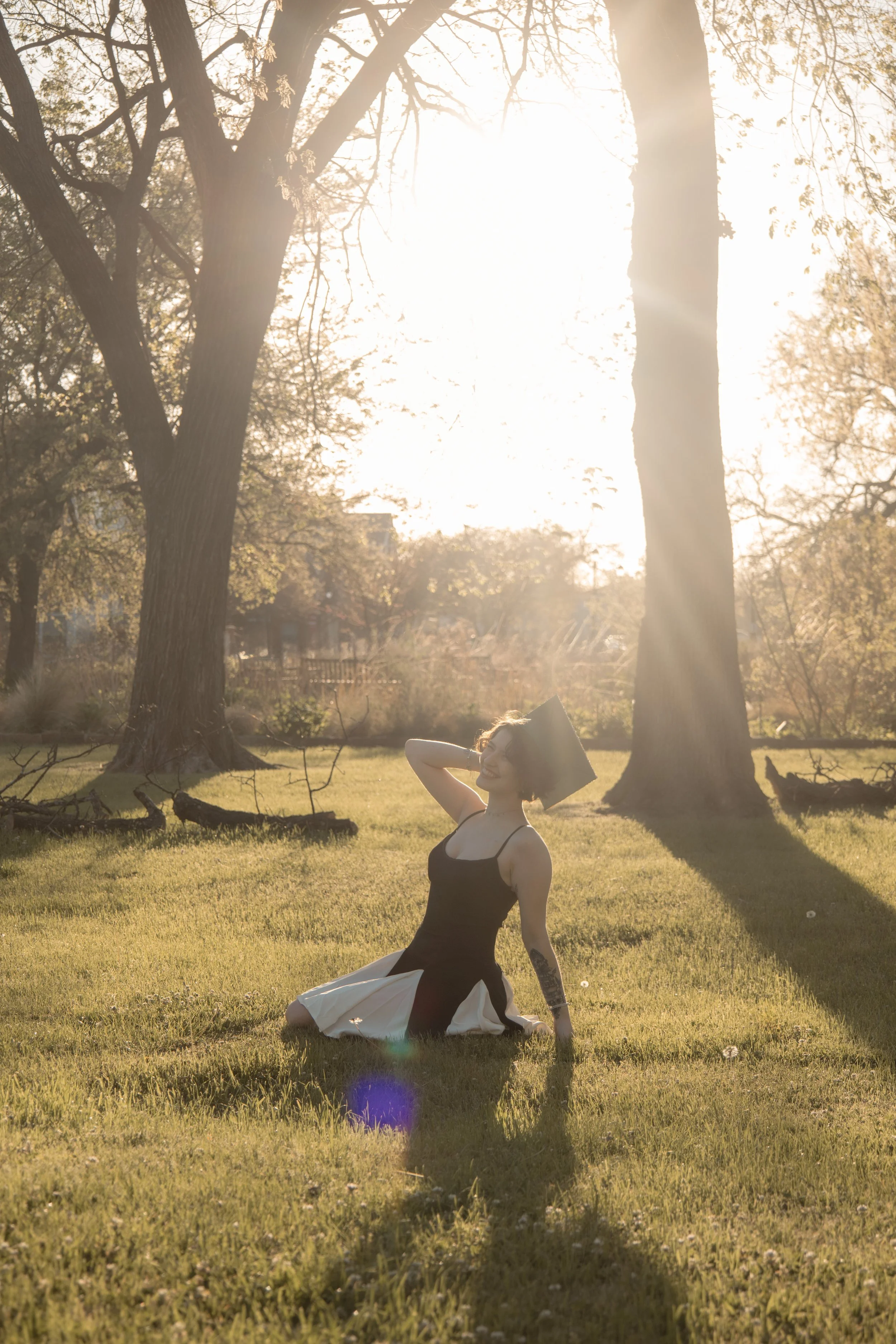 A woman in a black dress with a white skirt sits on the grass in a park, smiling with her eyes closed as sunlight filters through the trees in the background.