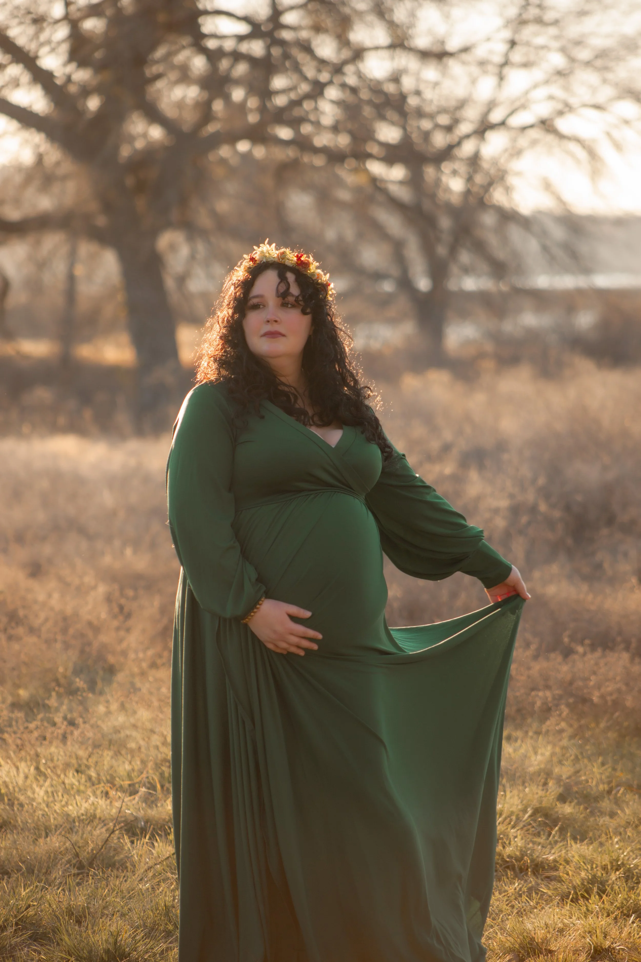 A pregnant woman with curly dark hair wearing a long green dress in a field at sunset, holding her dress with one hand and resting her other hand on her belly, with a tree in the background.