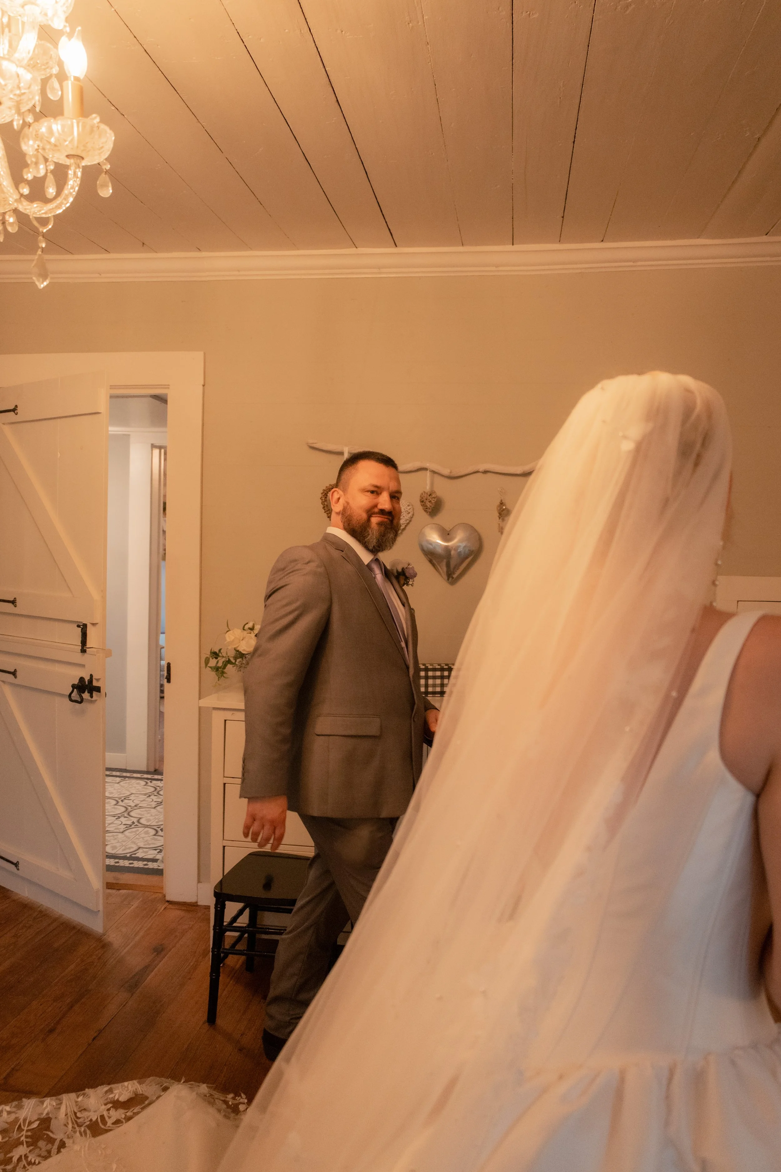 A man in a gray suit standing indoors near a wooden staircase, decorated with heart-shaped ornaments and silver balloons, with a chandelier and doorways visible in the background.