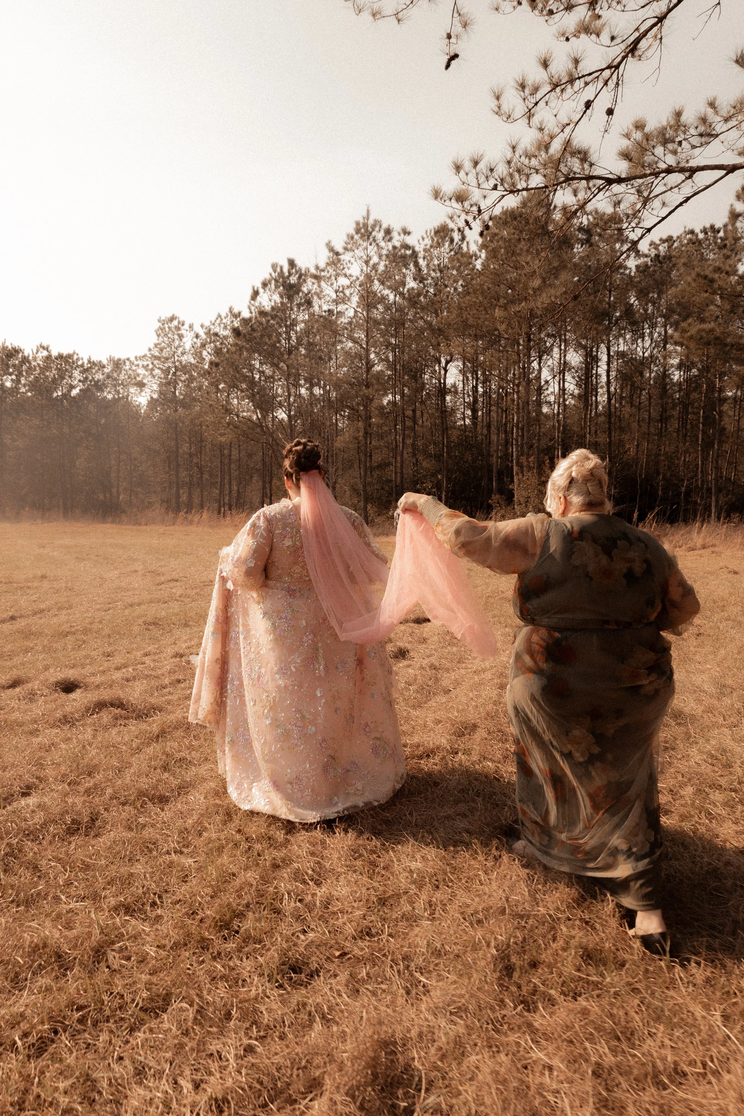 Two women walking through a field holding a pink veil, with one dressed in a floral gown and the other in a dark, patterned dress, and trees in the background under a clear sky.