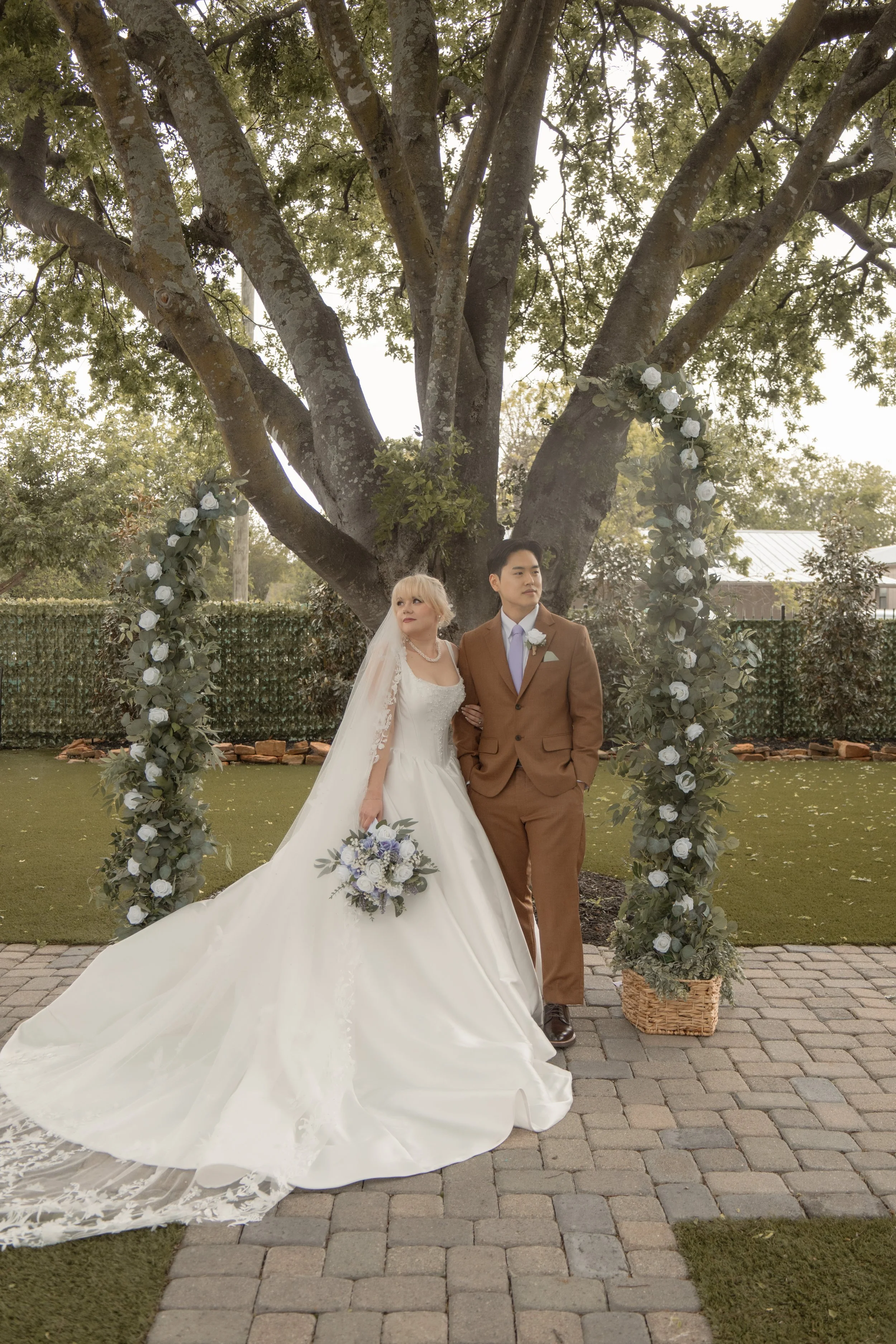 A bride and groom standing under a large tree decorated with white roses and greenery, posing for a wedding photo outdoors, with a stone-paved walkway and a wooden fence in the background.