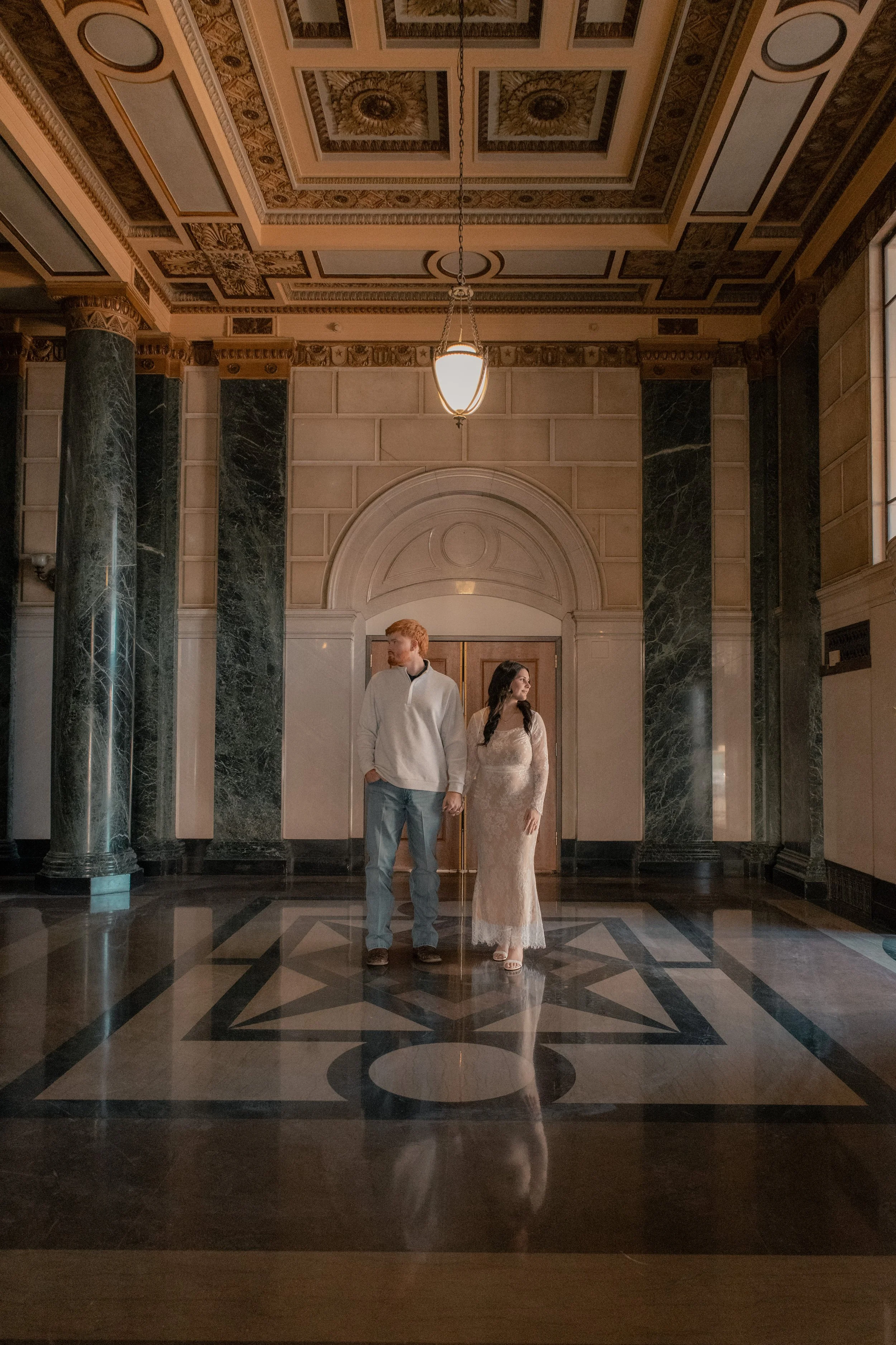 A man and woman standing hand in hand in a grand, ornate hall with marble pillars and a decorative ceiling ceiling.