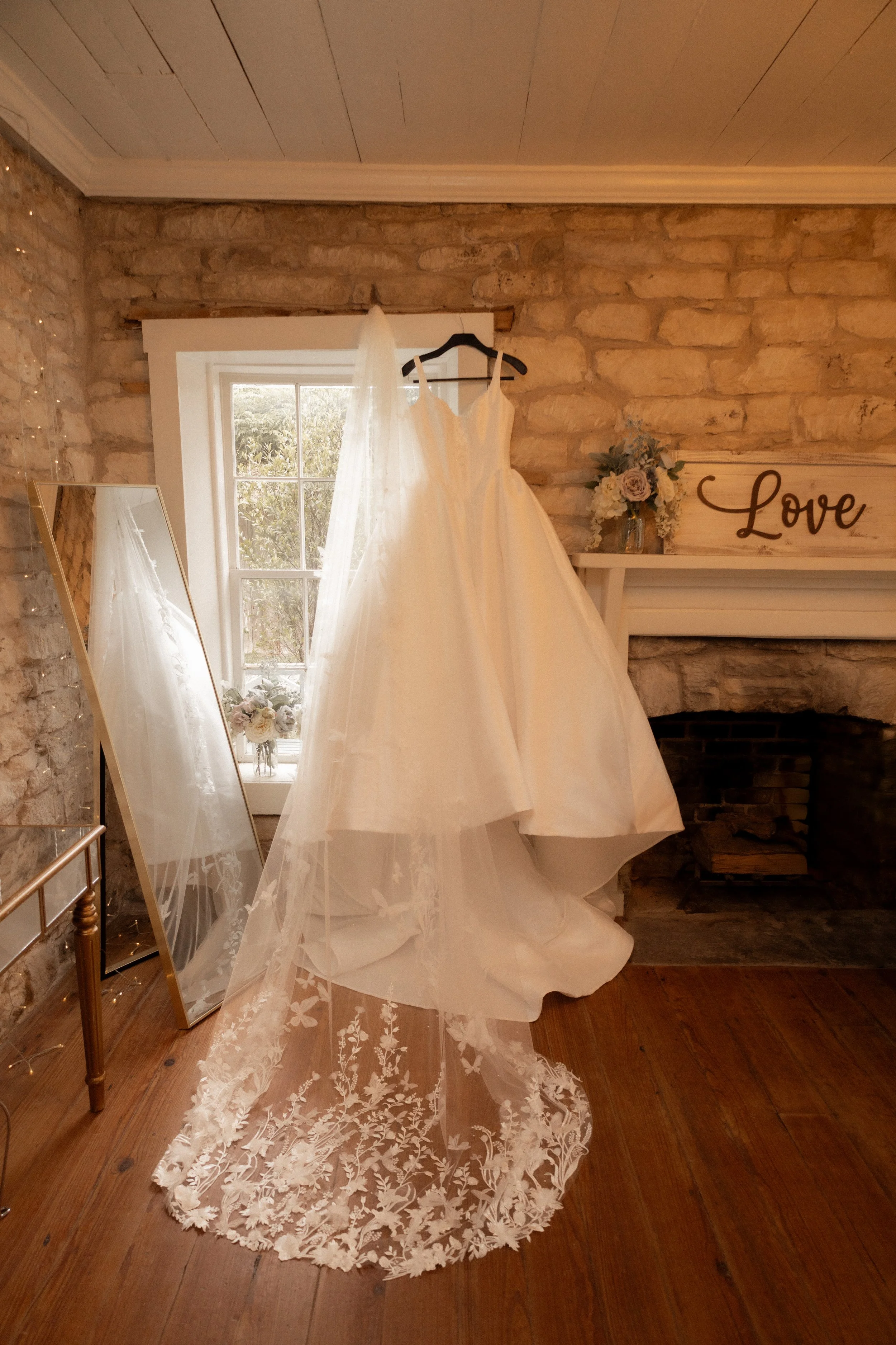 A wedding dress hangs on a black hanger in a rustic room with a stone chimney and wooden floors. The dress has a voluminous skirt and a delicate lace train extending onto the floor.